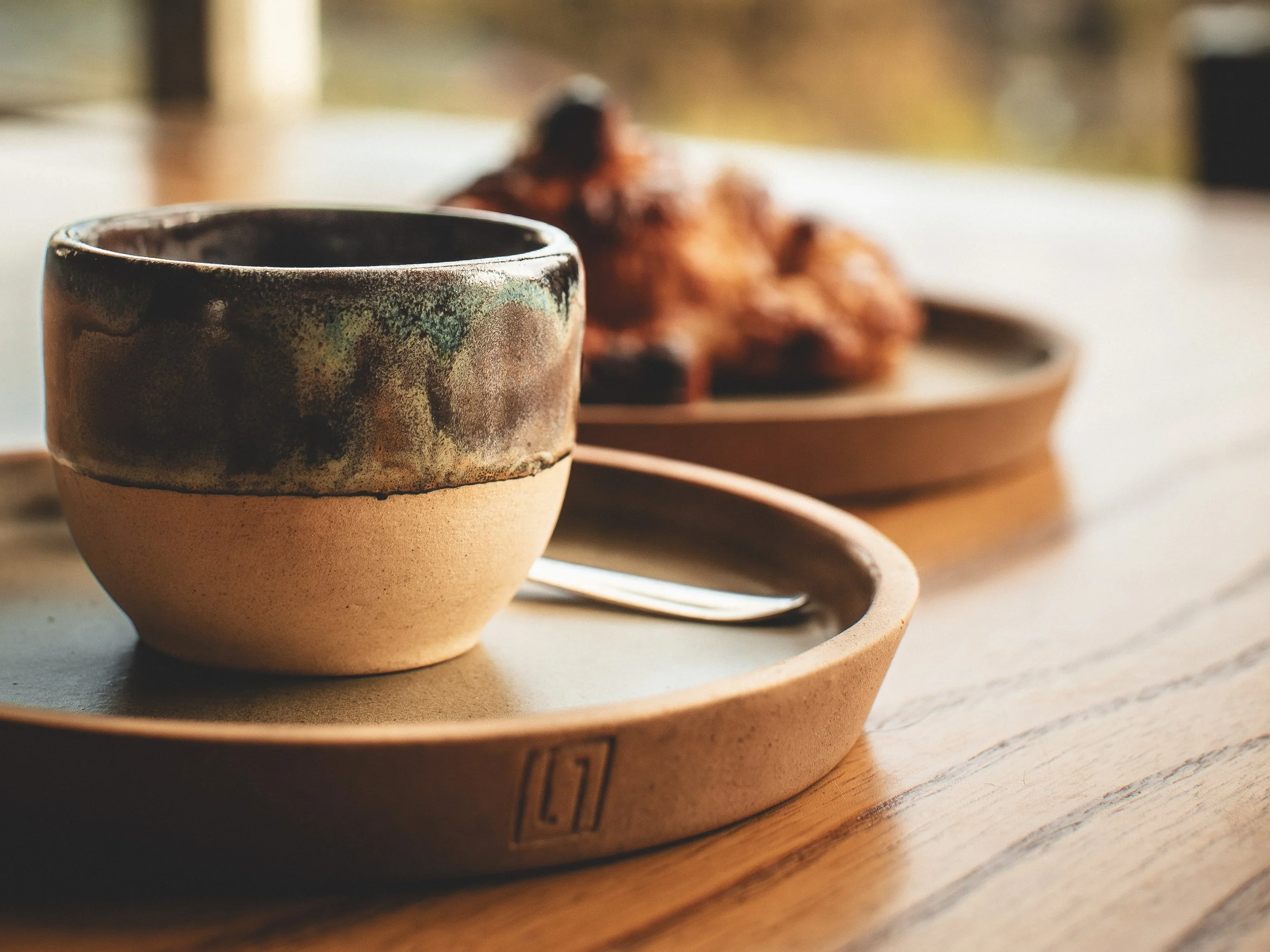 Taza de cerámica con esmalte decorativo en tonos marrón y verde, sobre una bandeja de cerámica en una mesa de madera, con comida al fondo.