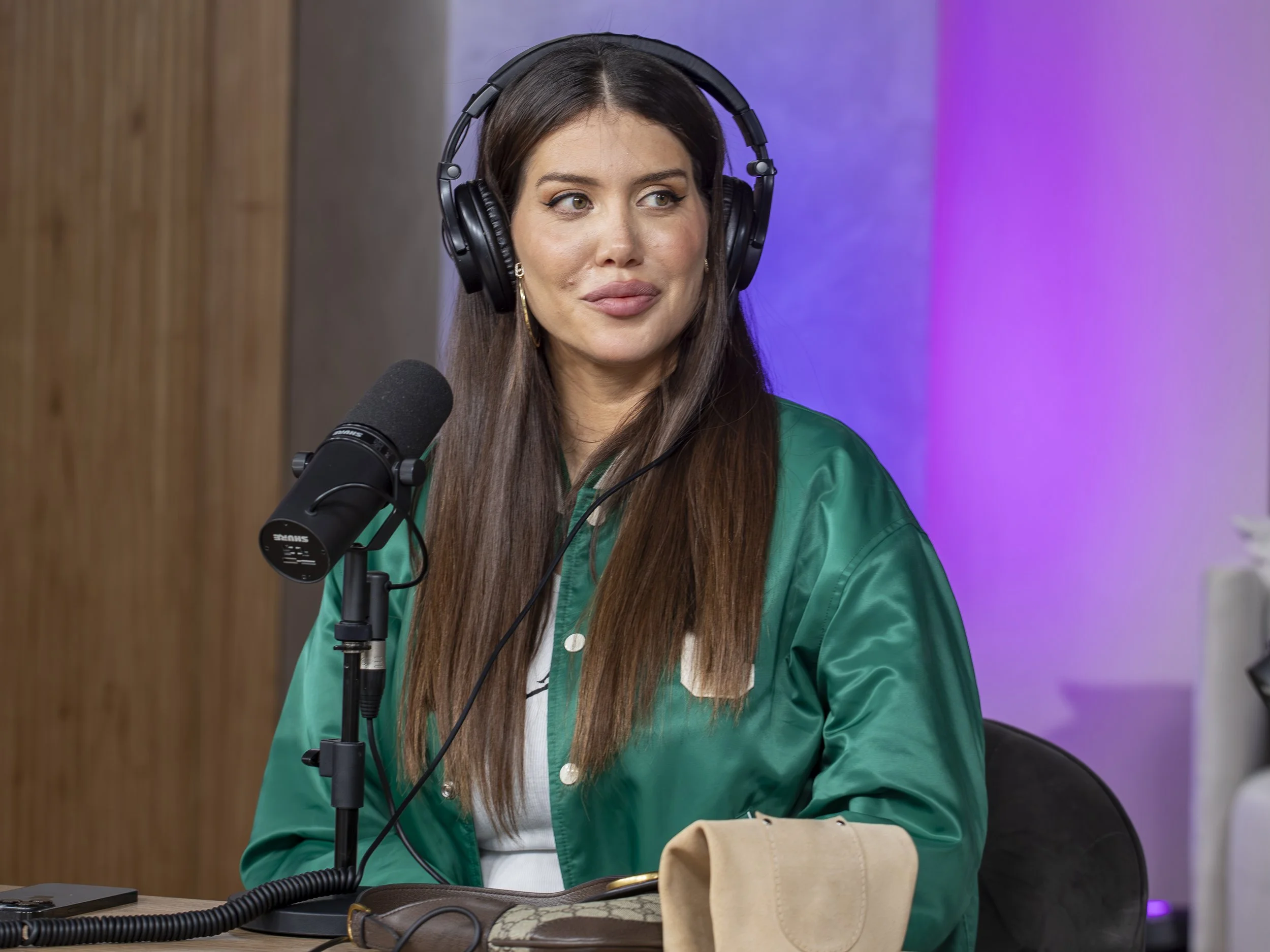 Mujer con cabello largo y suelto usando audífonos grandes y camiseta verde, hablando frente a un micrófono en un estudio de grabación con paredes de color morado y madera.