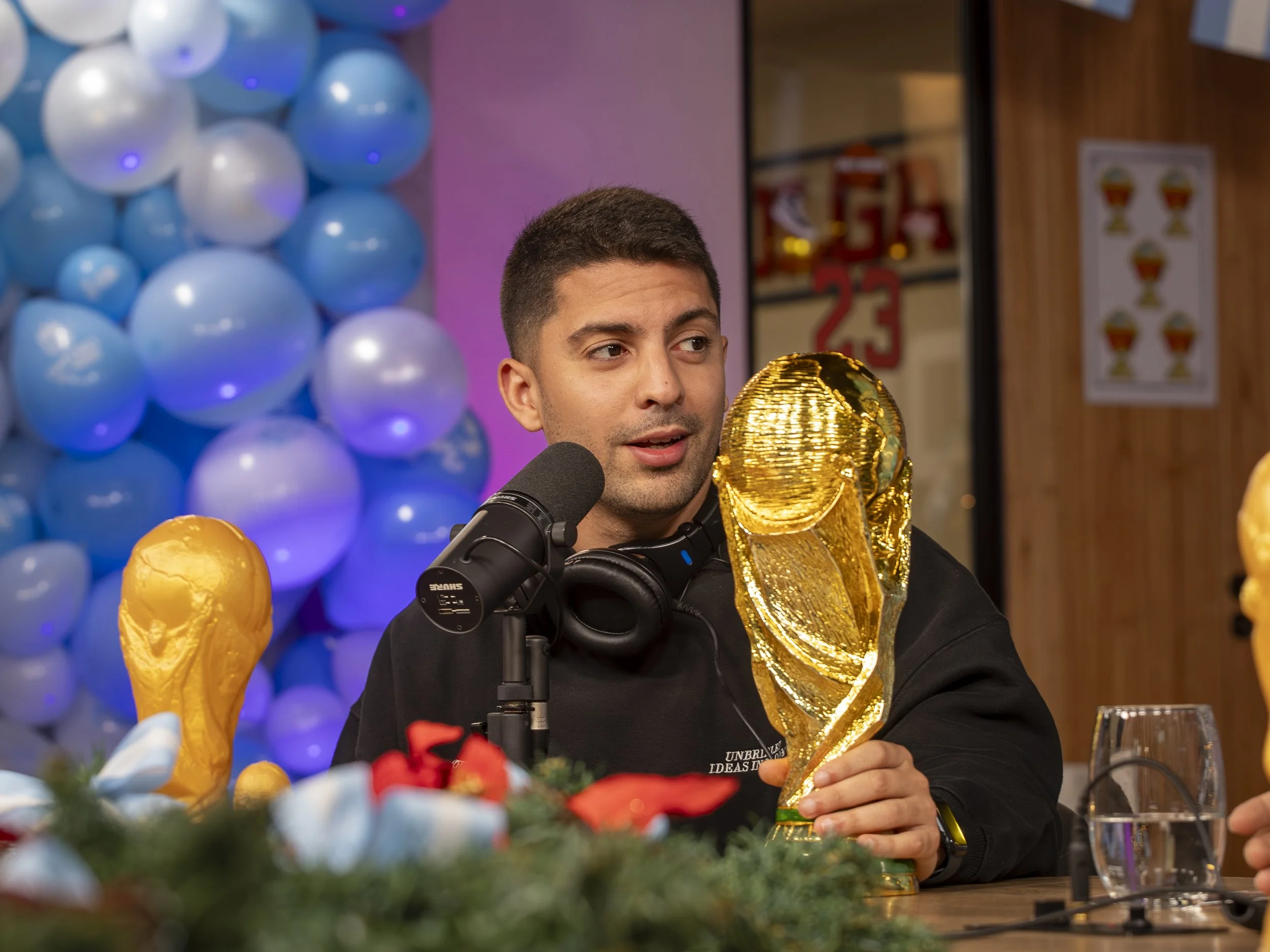 Un hombre en un estudio decorado con globos y símbolos deportivos, sosteniendo un trofeo dorado de la Copa del Mundo de la FIFA.