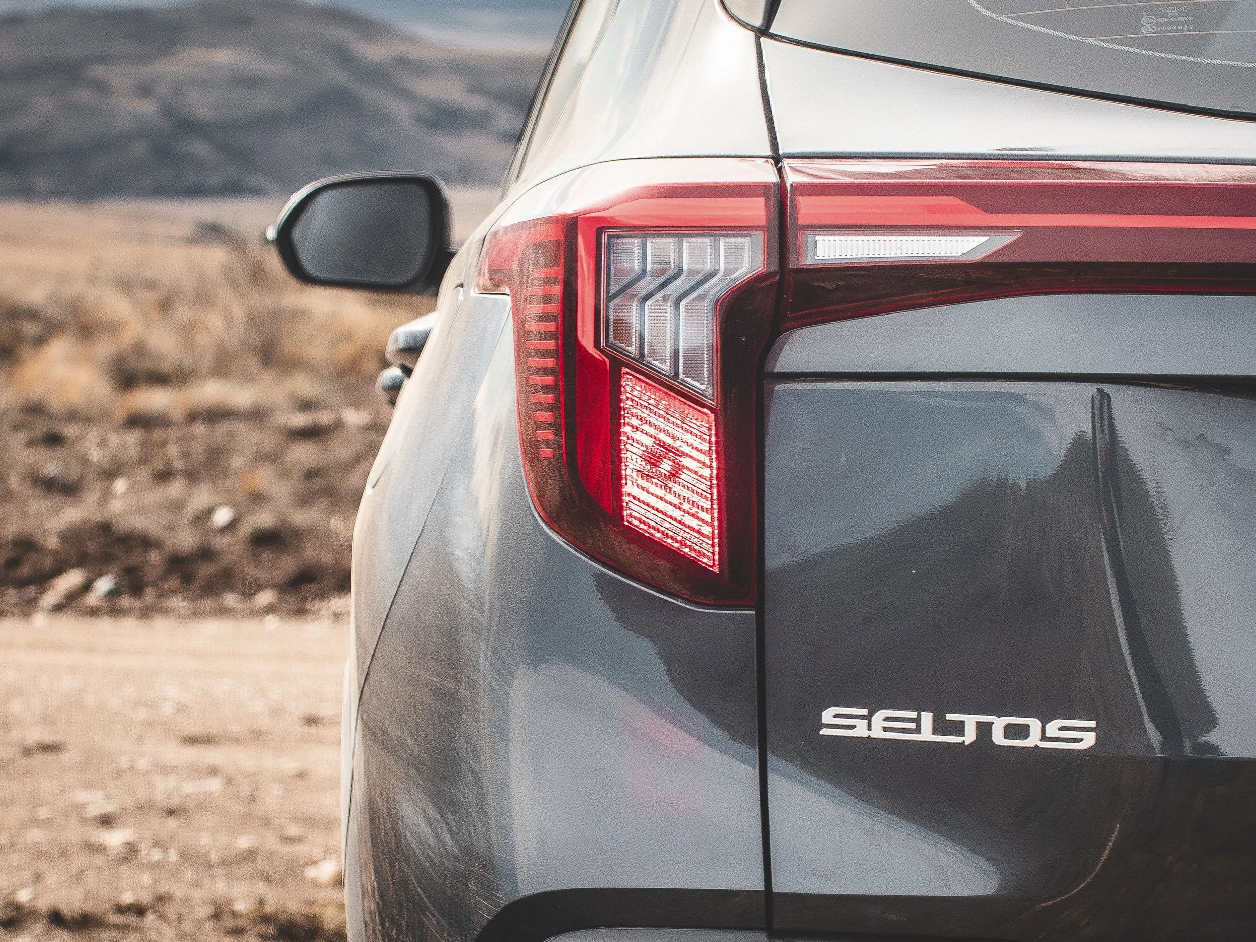 Parte trasera de un automóvil gris con la matrícula 'SALOS', mostrando la luz trasera y el espejo lateral, en un paisaje de tierra árida.