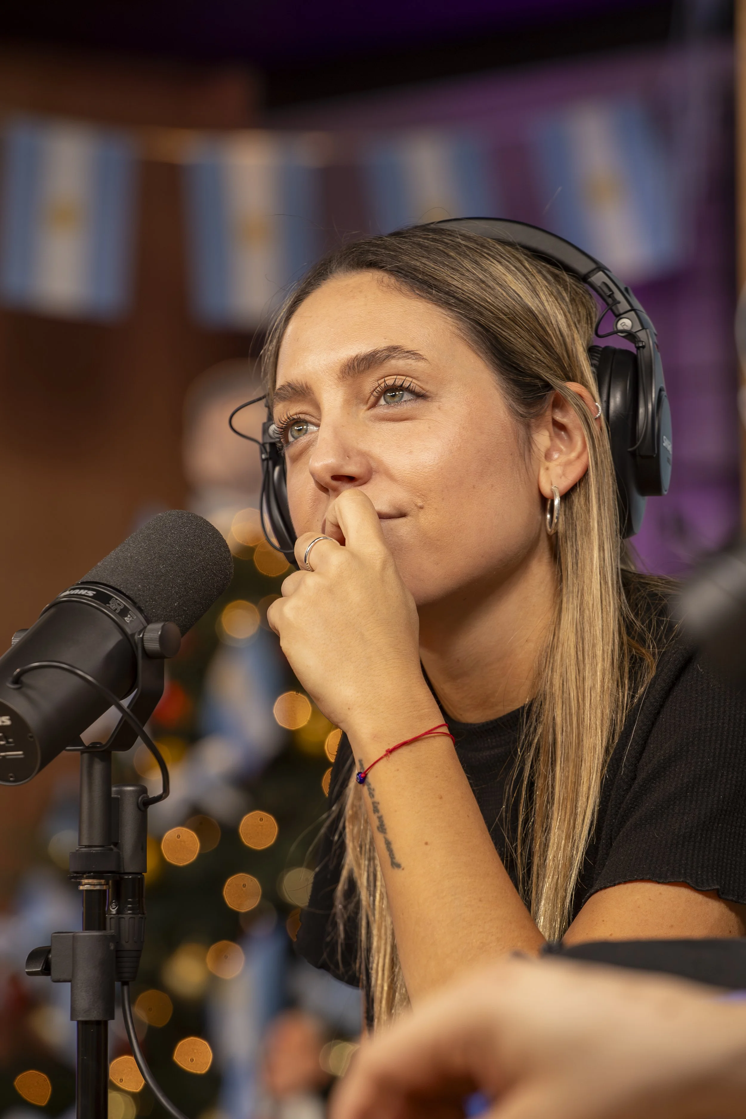 Mujer con auriculares frente a micrófono en un estudio, con fondo de luces difusas y decoraciones navideñas.