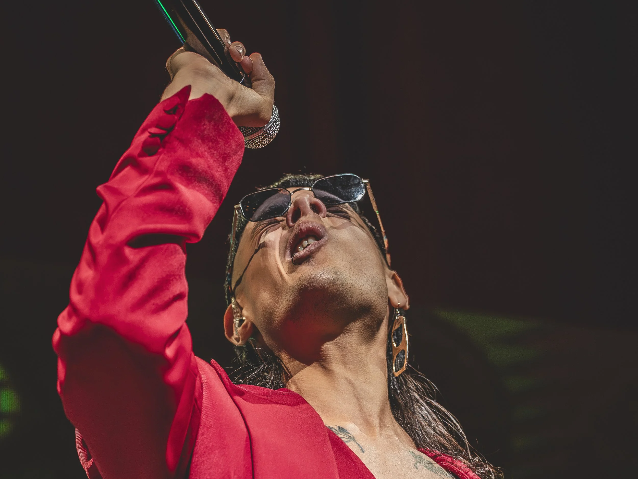 Persona cantando con gafas de sol, pelo oscuro, vestido rojo, en un escenario oscuro.