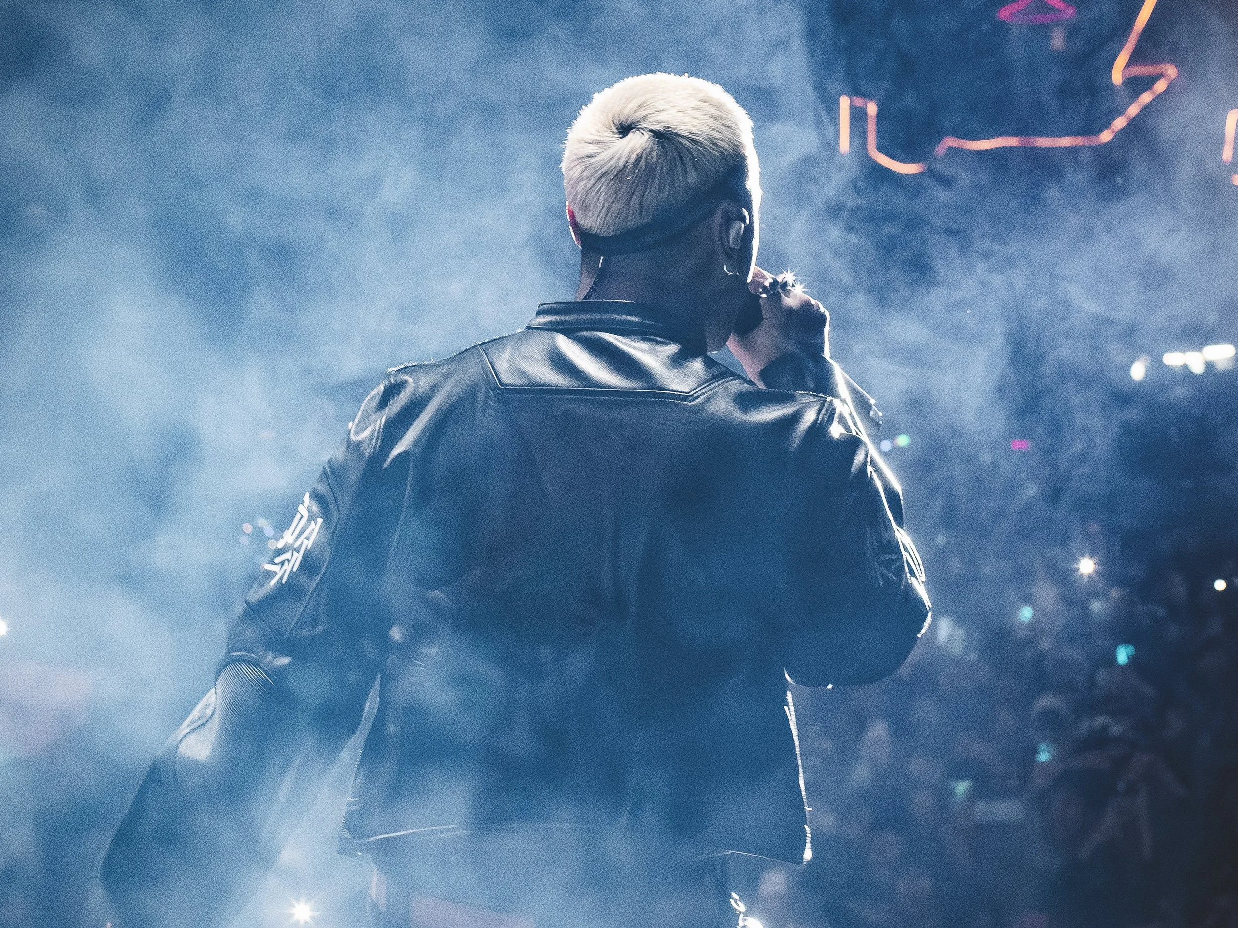 Persona con cabello rubio platinado, vista de espaldas, con chaqueta de cuero negro, en un escenario con humo y luces de neón.