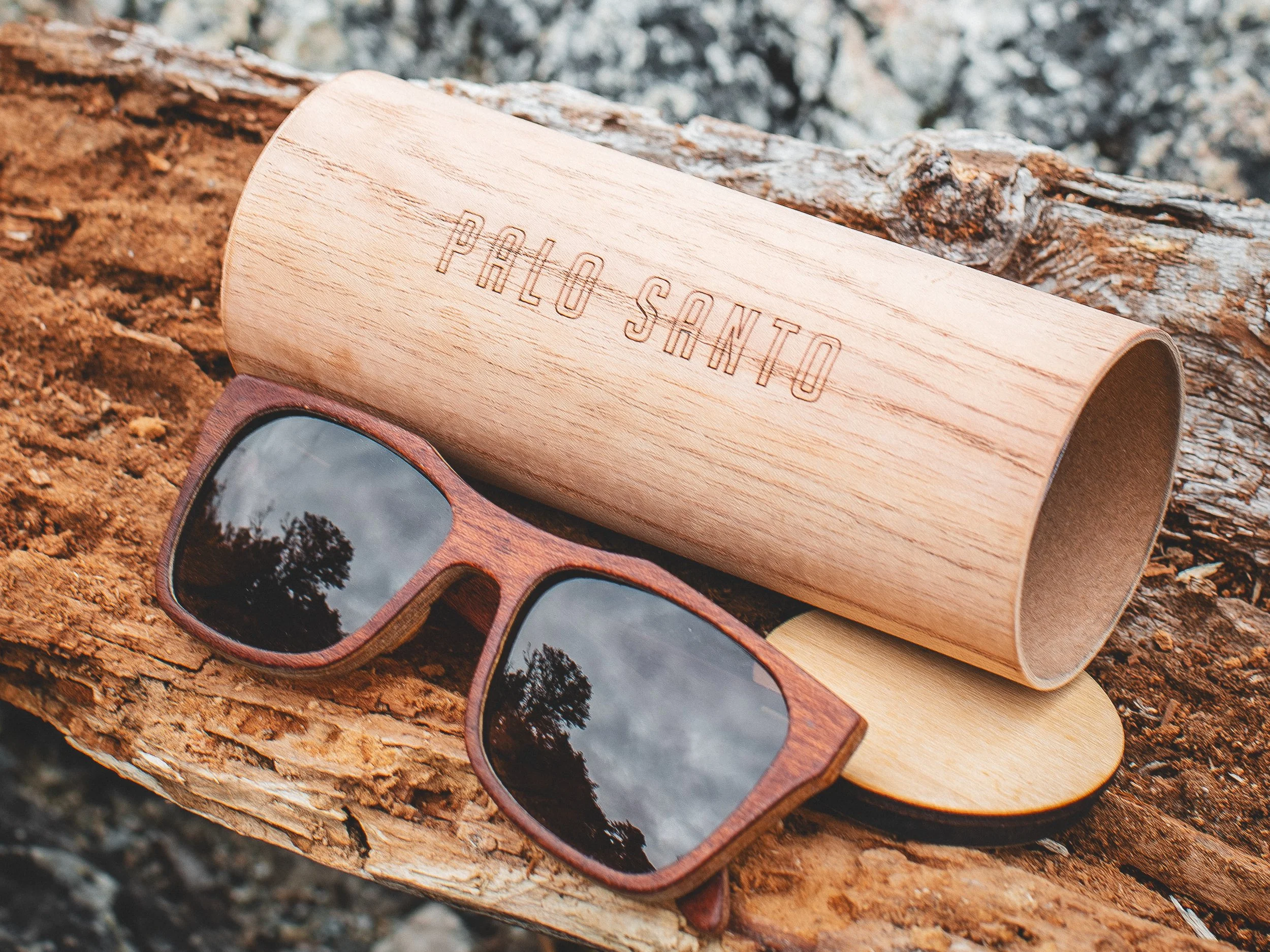 Un par de gafas de sol de madera junto a una caja de madera grabada con la inscripción 'Palo Santo', ubicadas sobre un tronco de árbol en un entorno natural.