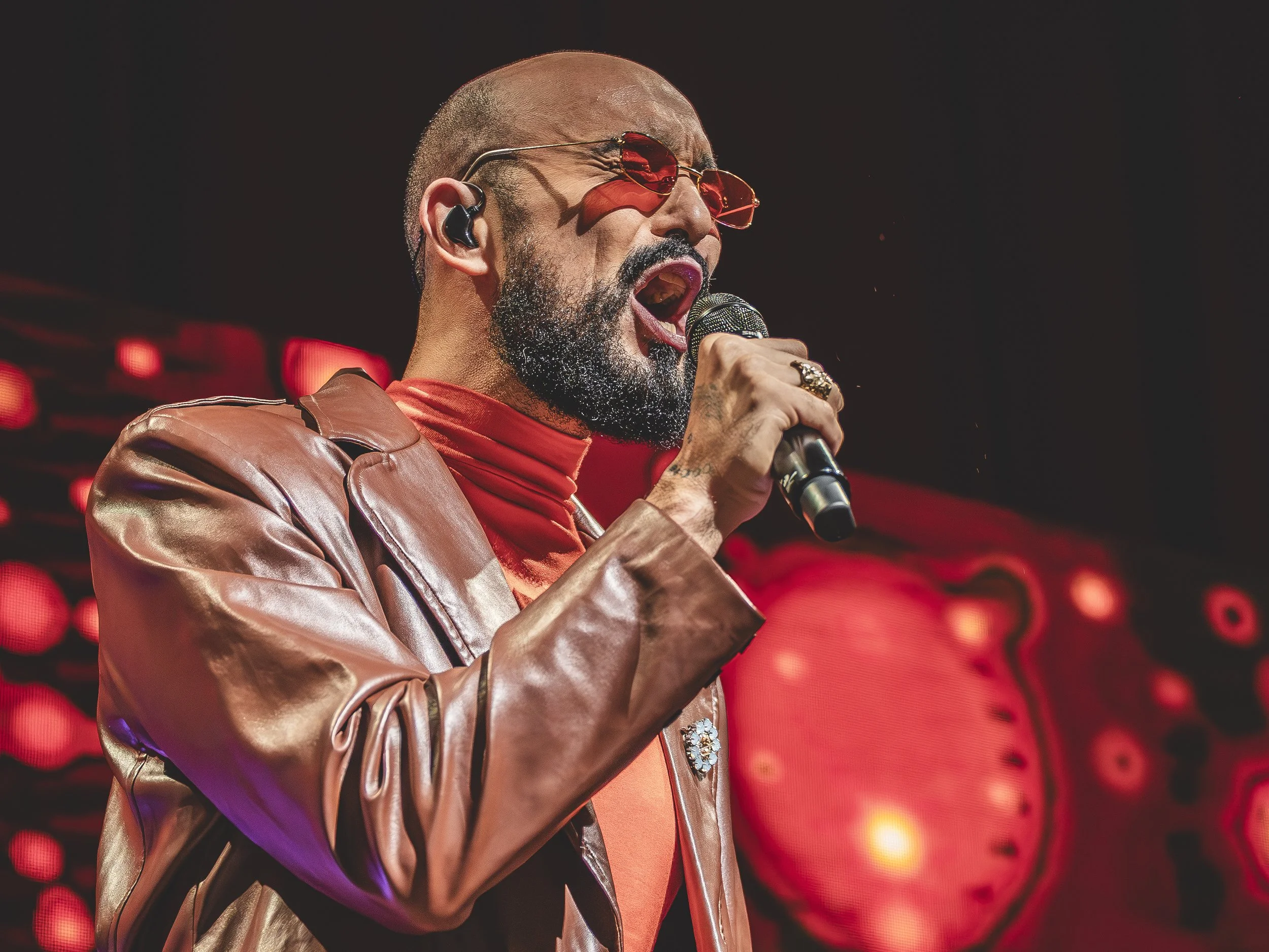 Hombre cantante con barba, gafas de sol rojas, vestimenta de cuero marrón, cantando en concierto con micrófono en mano y fondo con luces rojas y una pantalla con diseño abstracto.