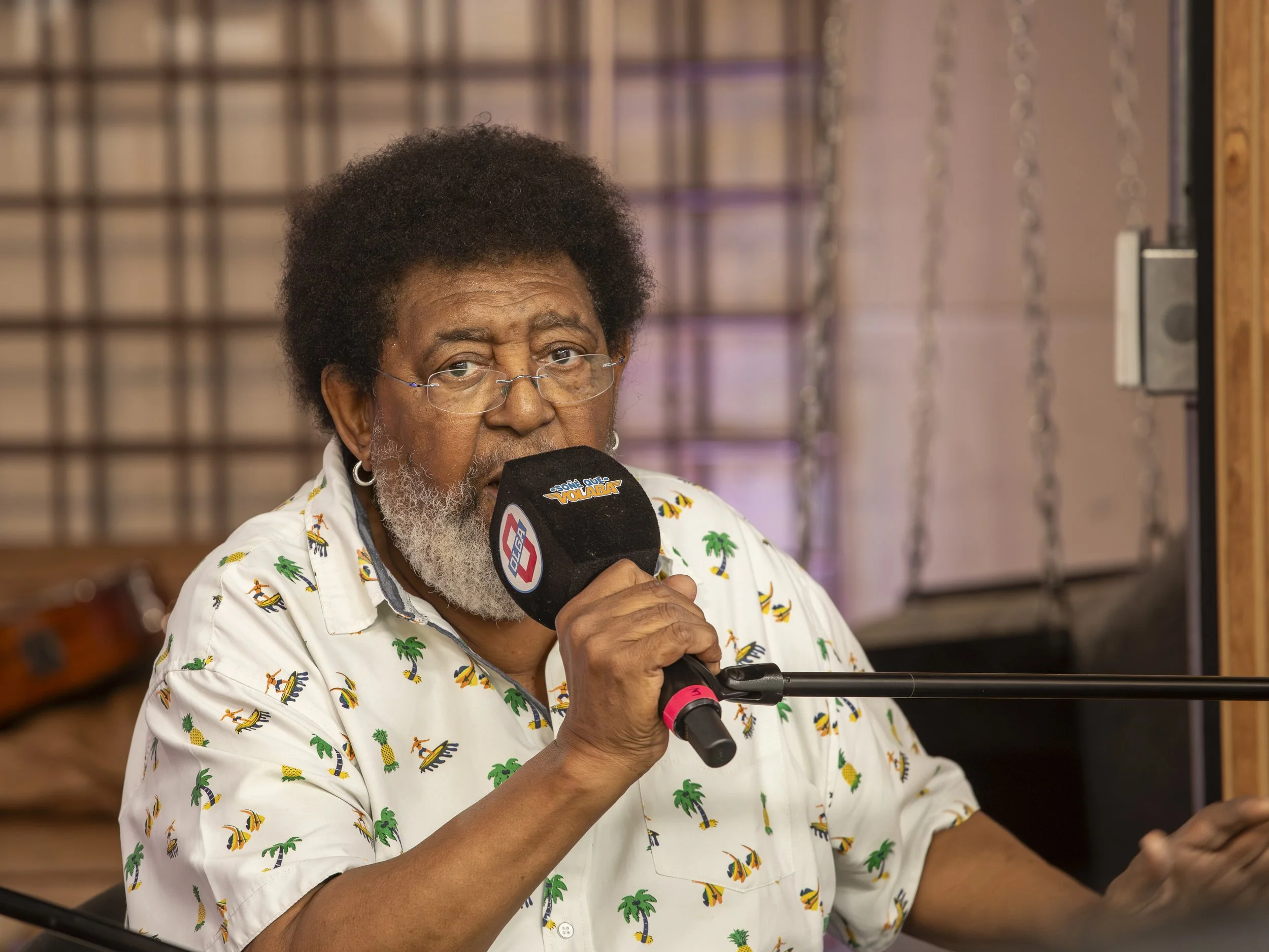 Hombre de cabello rizado, barba y gafas, sosteniendo un micrófono con logotipo. Lleva una camisa de colores claro con estampado de palmeras y elementos tropicales, en un entorno interior con decoraciones de madera y paneles.