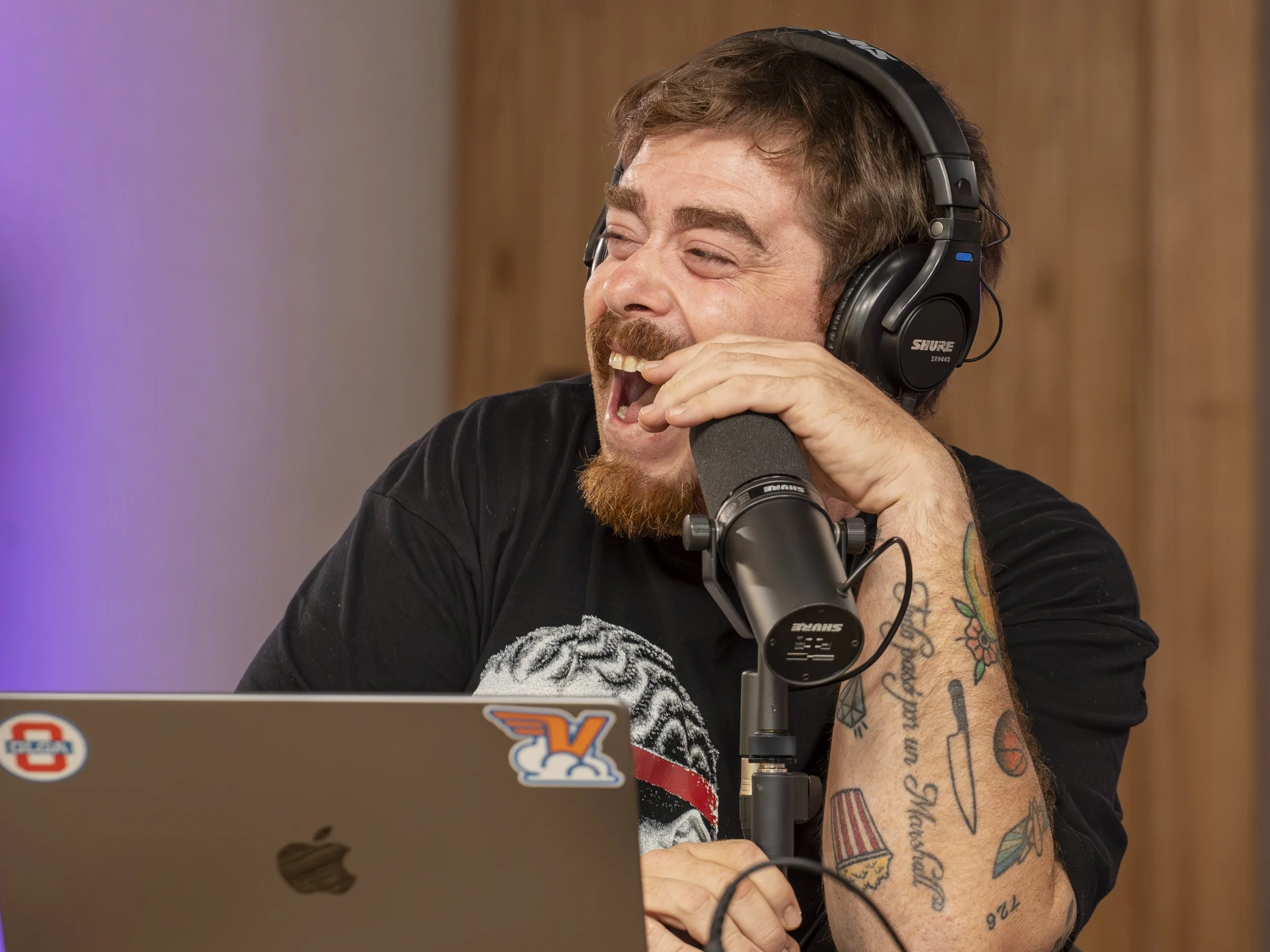 Hombre con barba y tatuajes riendo, lleva audífonos y habla en un micrófono en un estudio.