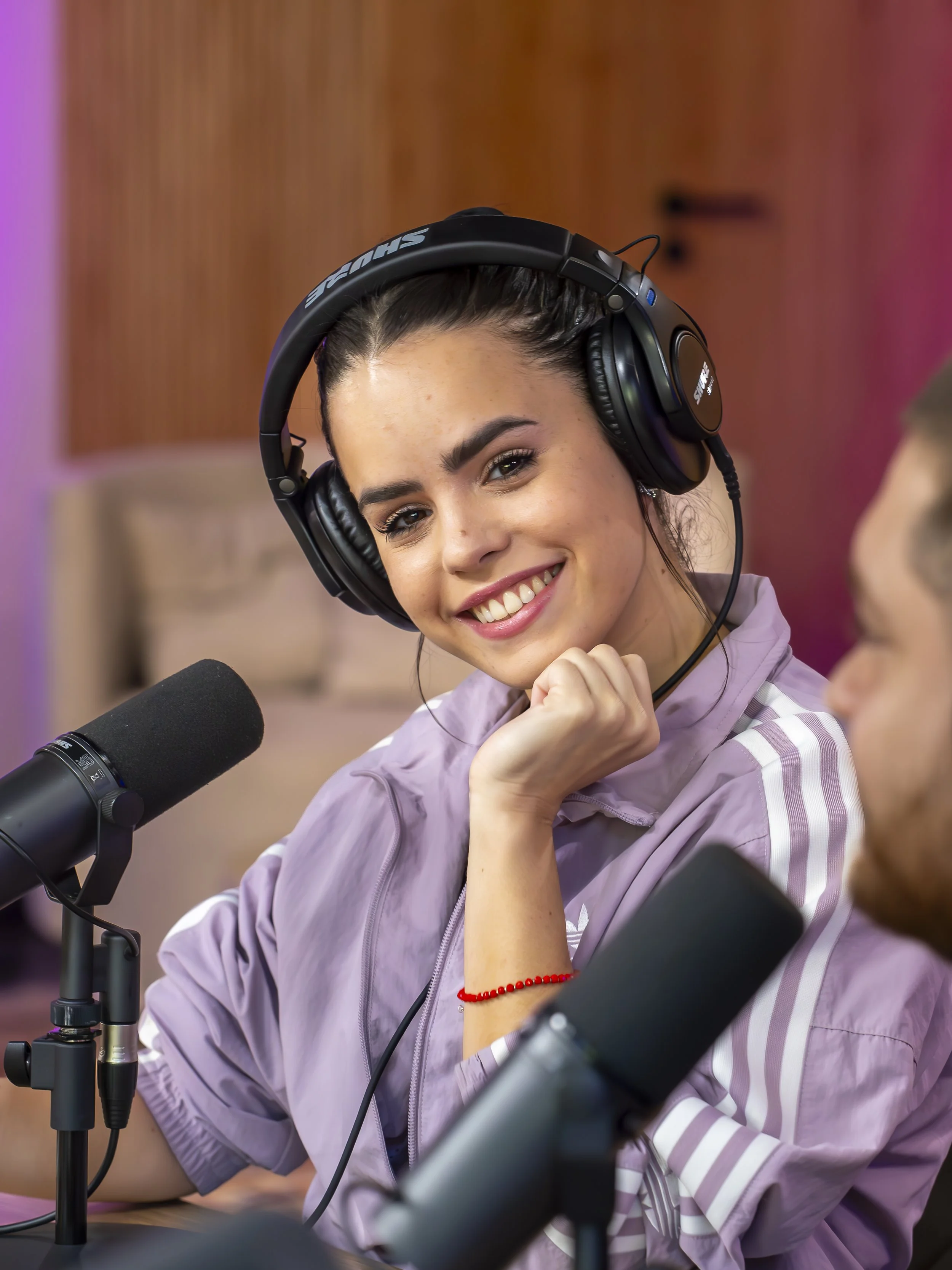 Joven mujer usando audífonos grandes en un estudio de grabación, sonriendo y hablando en micrófono, con otro hombre en la conversación.