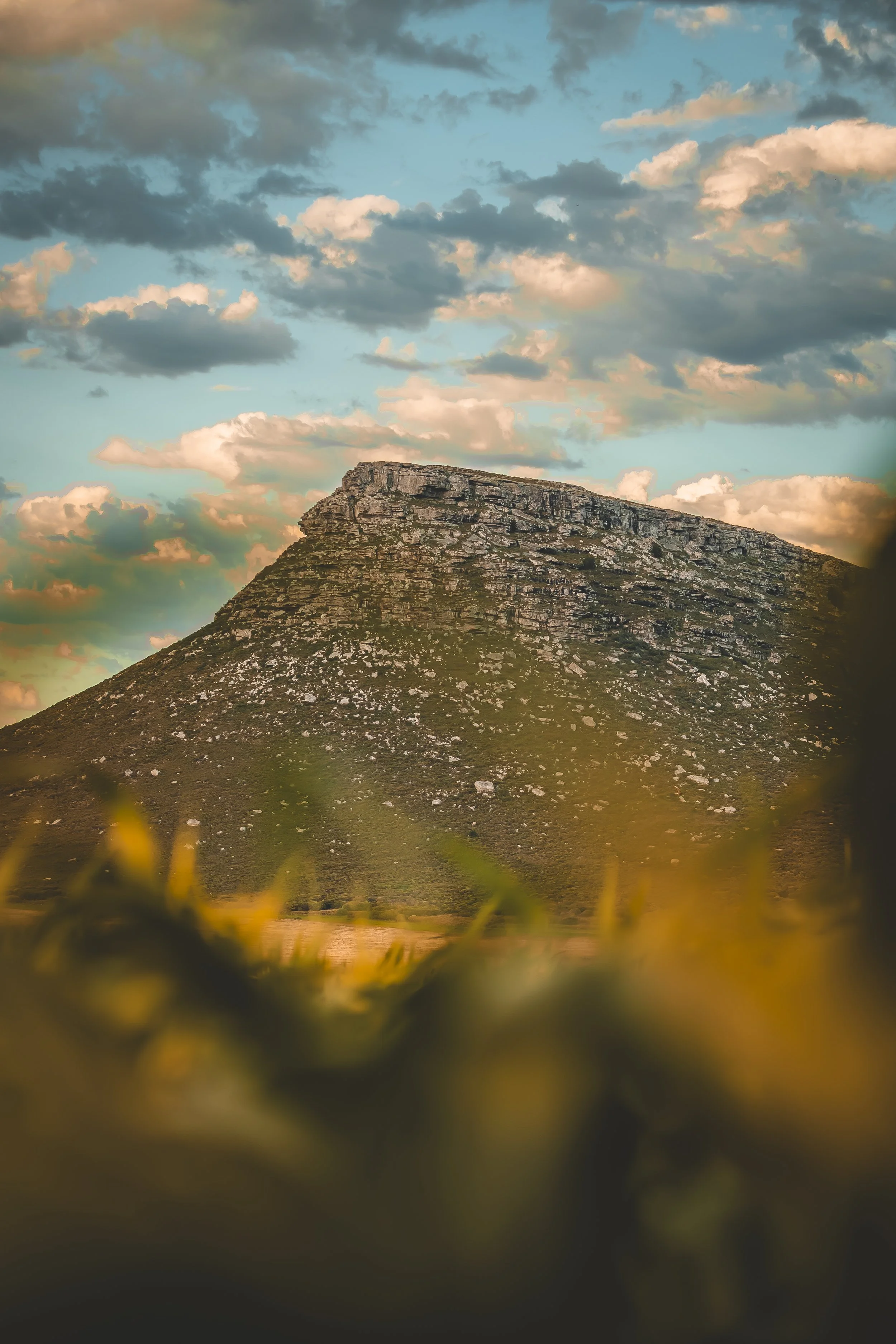 Vista de una montaña con forma de meseta, rodeada de naturaleza, con cielo nublado y atardecer.