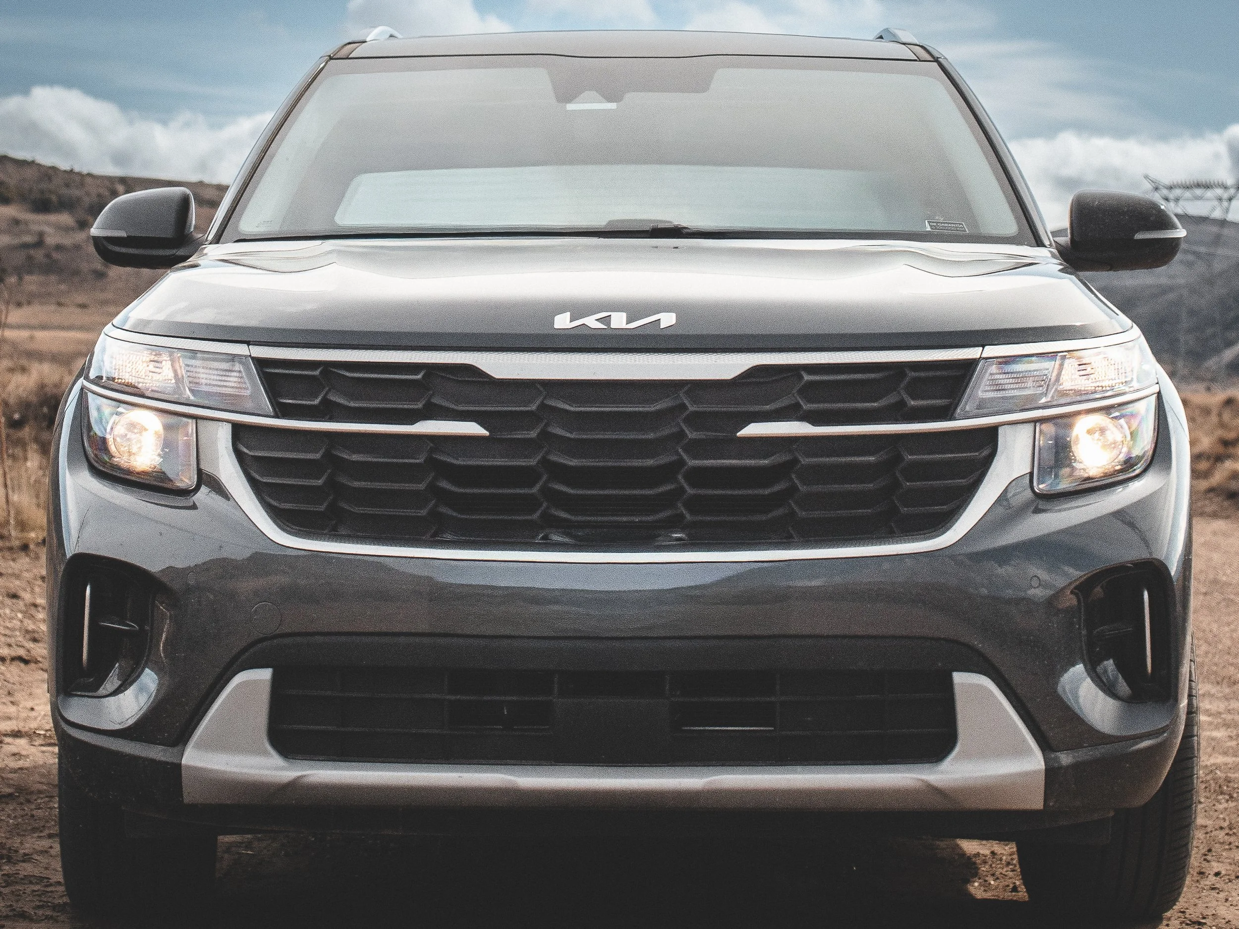 Coche Kia gris en un terreno arenoso con cielo nublado de fondo.