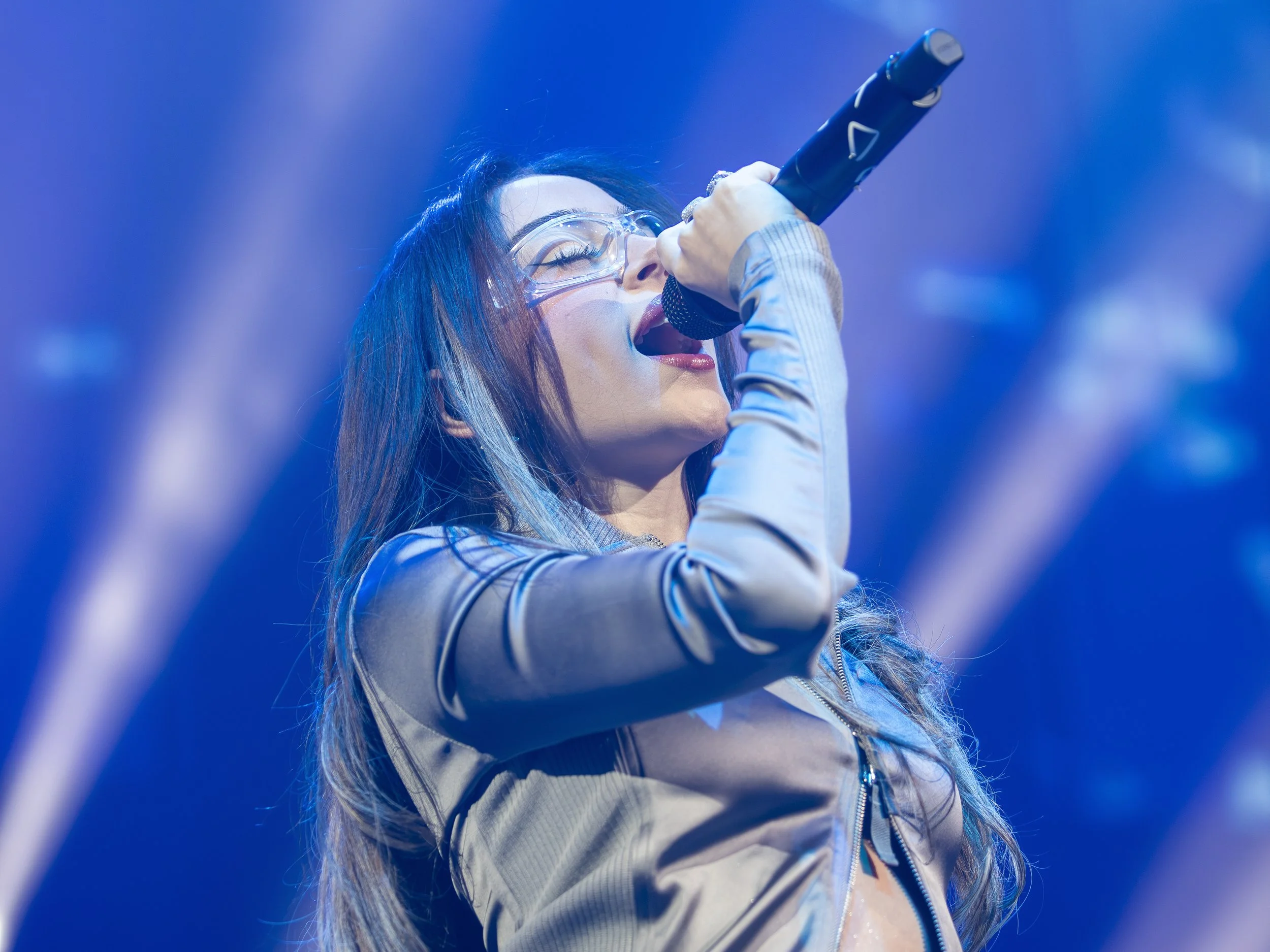 Una mujer cantante con gafas y cabello largo, entonando en un micrófono en un escenario con iluminación azul.