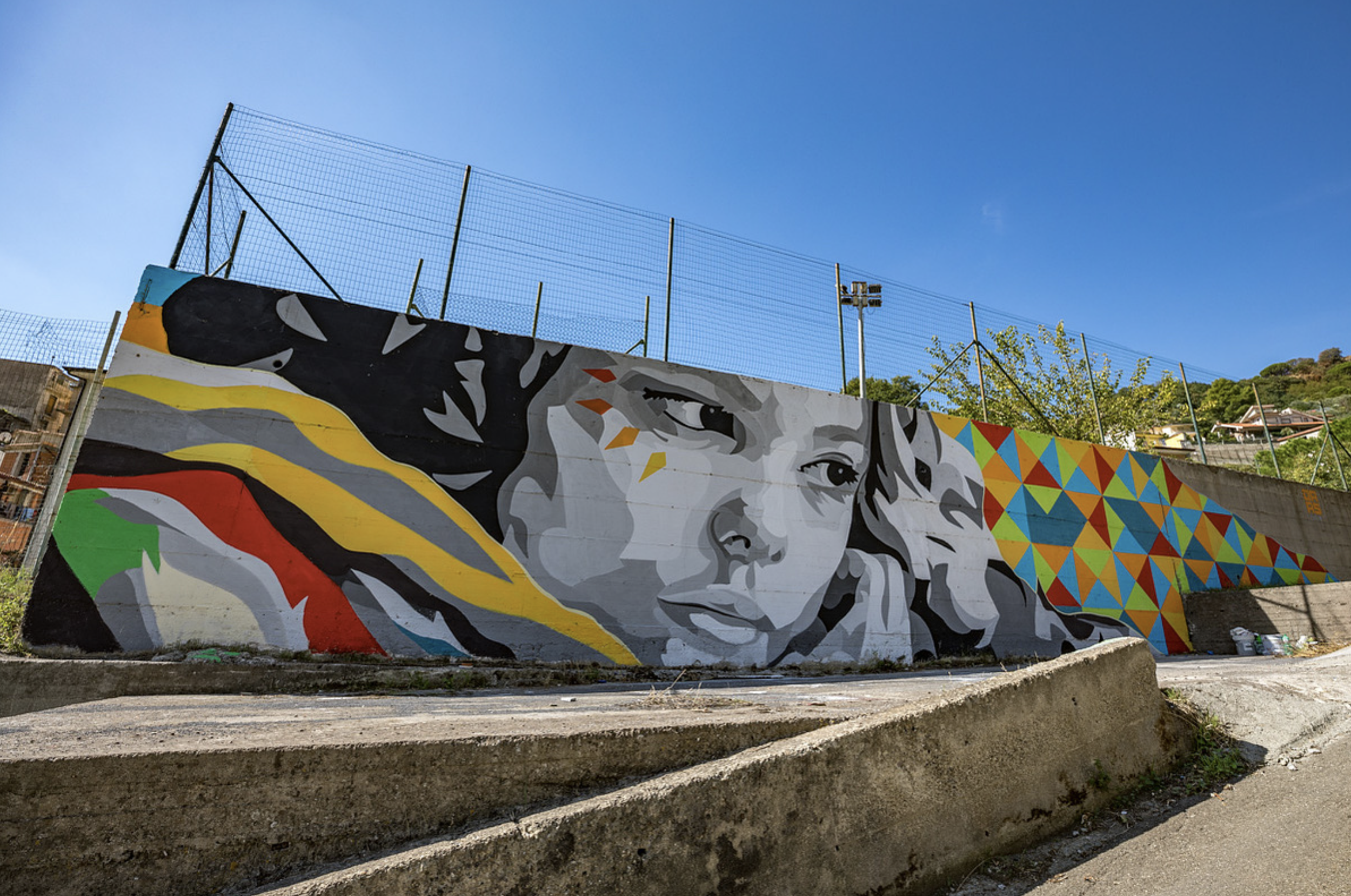 Work by the artist Colorful mural on a concrete wall depicts a grayscale portrait of a young girl with rainbow-like streaks on her face, next to a vibrant geometric pattern of triangles in red, blue, green, yellow, and orange, under a clear blue sky.