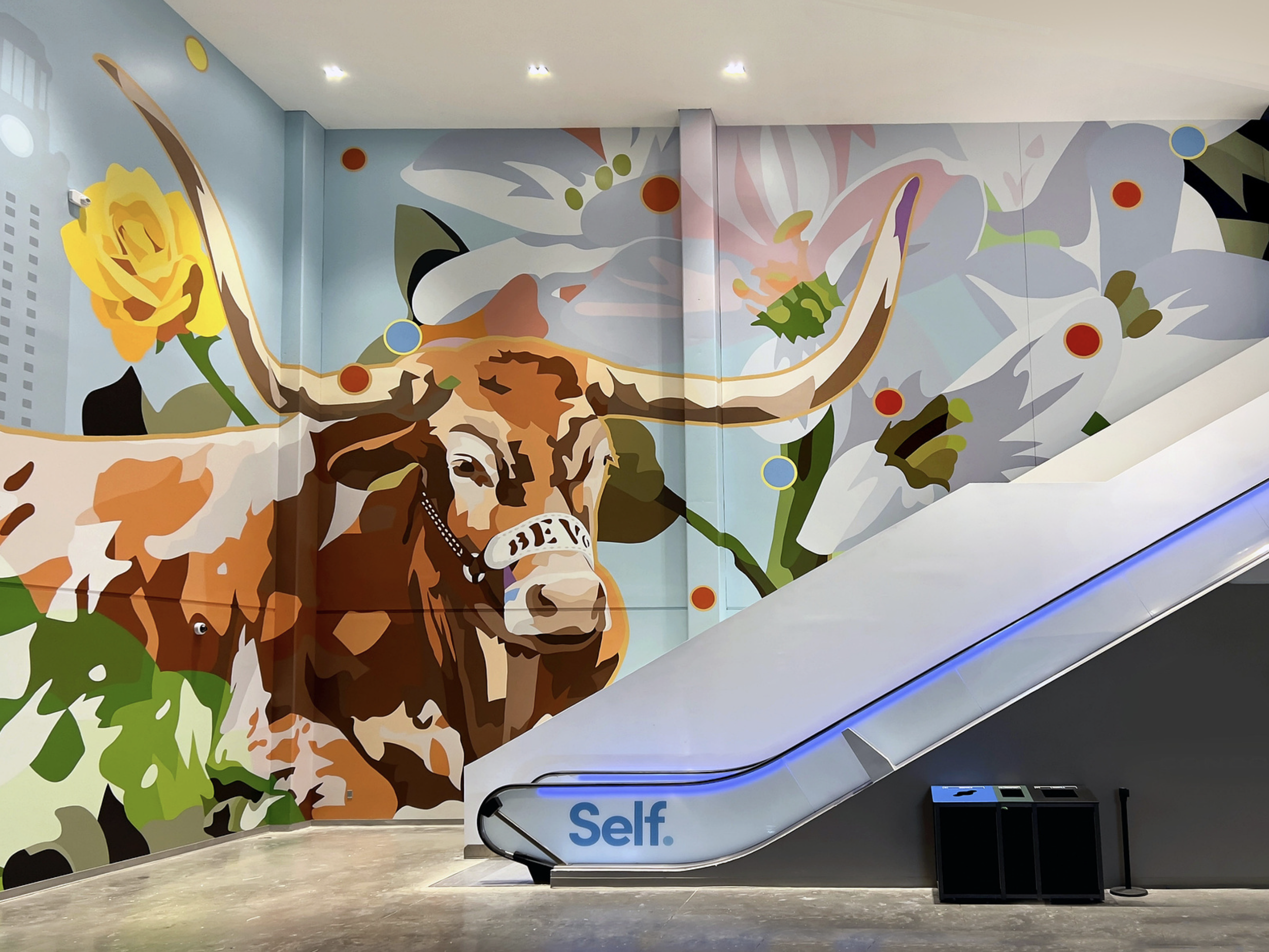 Work by the artist DAAS, Colorful mural of a highland cow with large horns, surrounded by large white flowers on a wall next to an escalator in a modern indoor space.