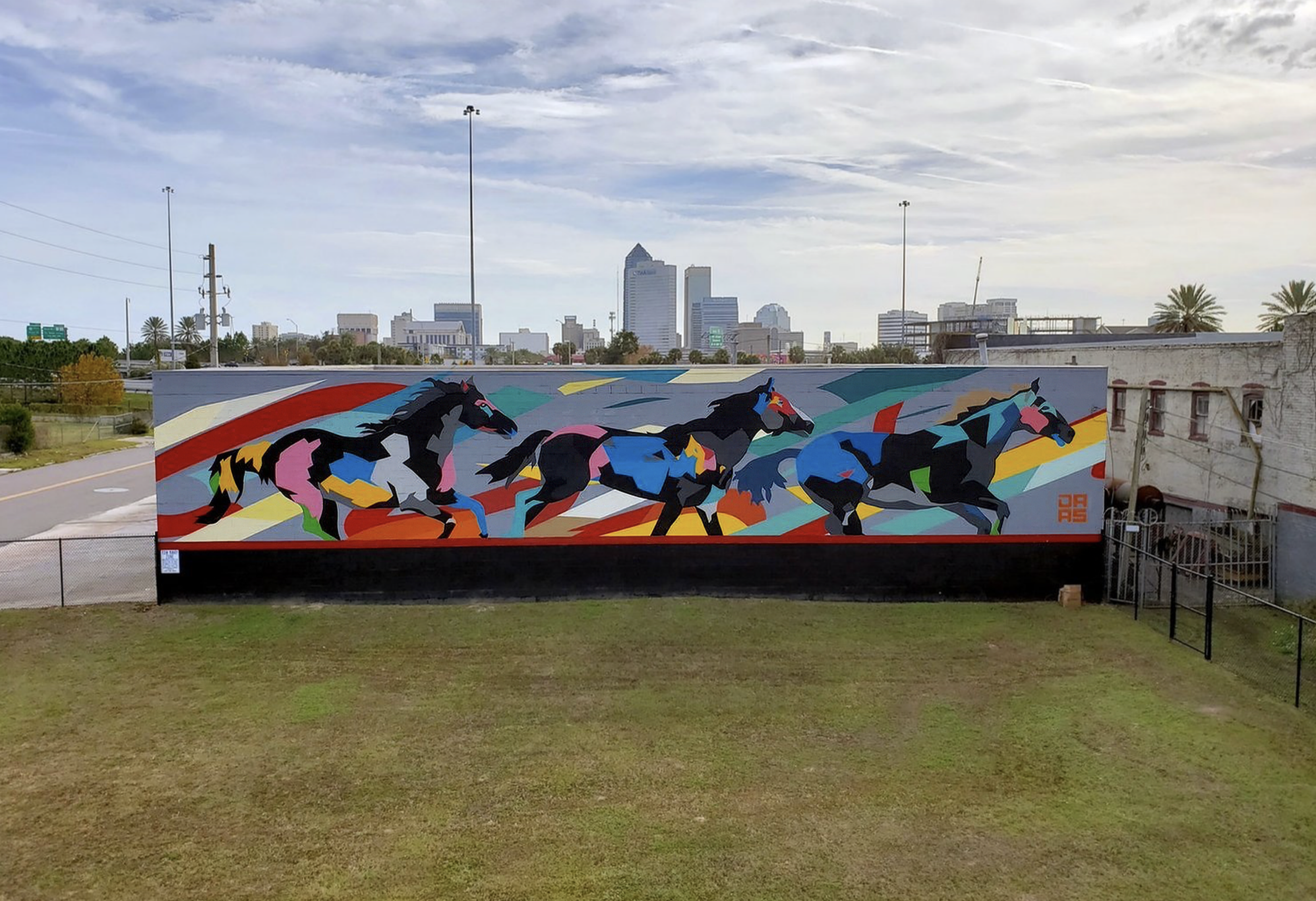 Work by the artist DAAS, Colorful mural of four racehorses running, painted on a large wall with a city skyline in the background.