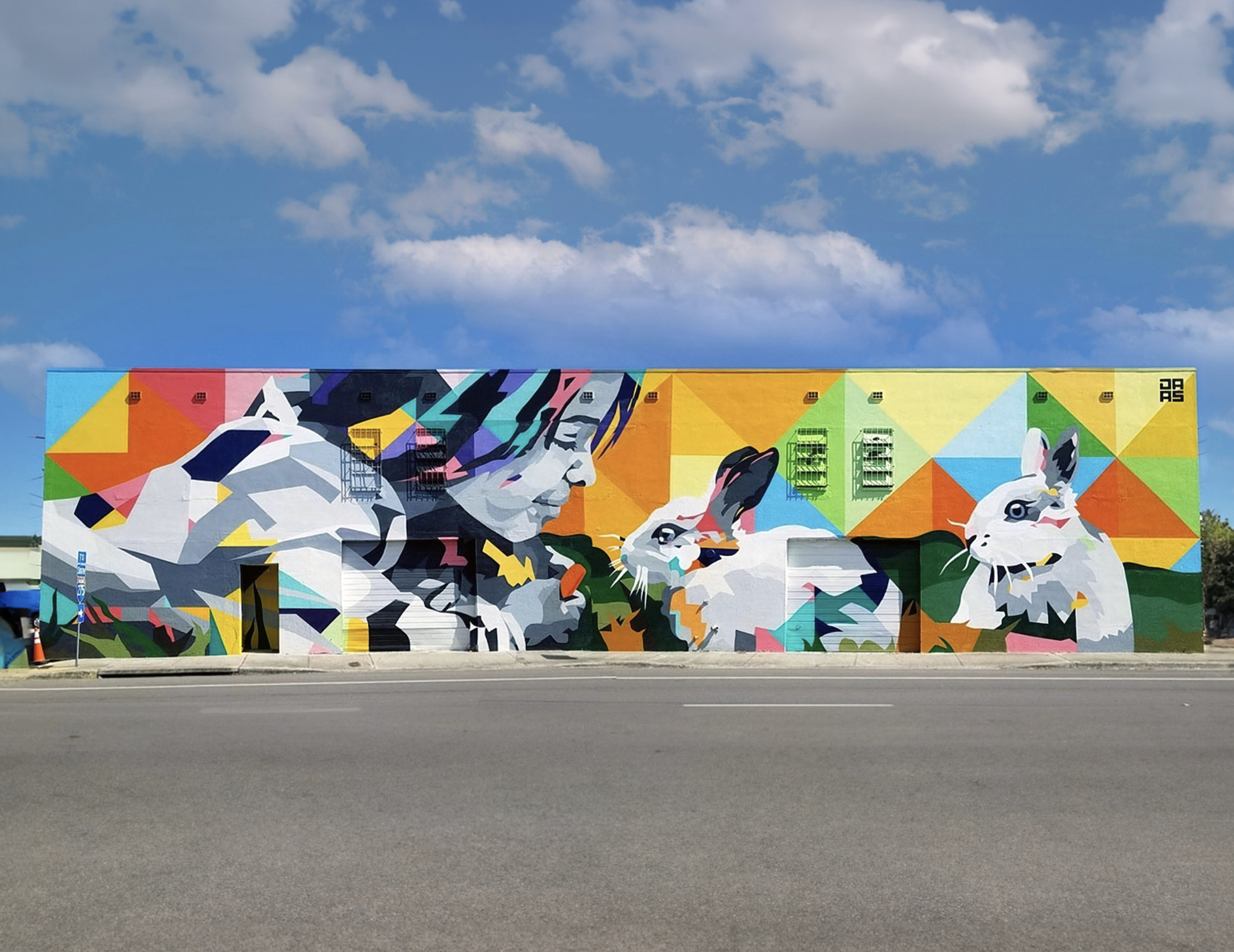 Work by the artist DAAS, Colorful mural of a woman and four rabbits on a building wall, with a blue sky and clouds in the background.
