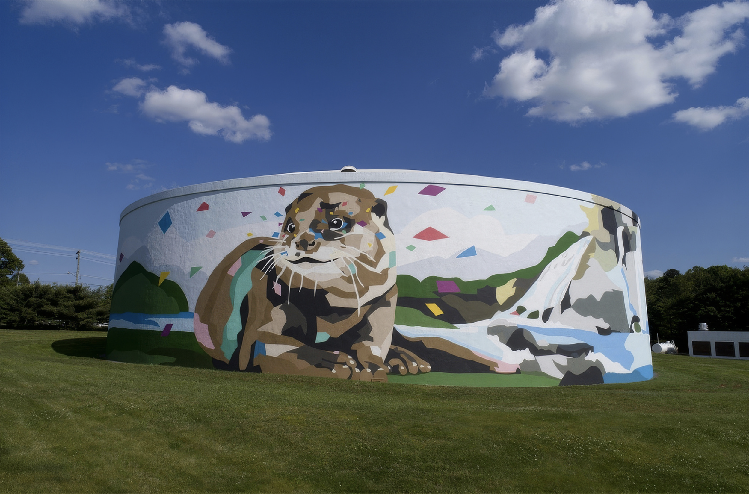 Work by the artist DAAS, Large round water tank with a mural of a mountain lion, a waterfall, and a mountain landscape, decorated with colorful confetti shapes, under a blue sky with clouds.