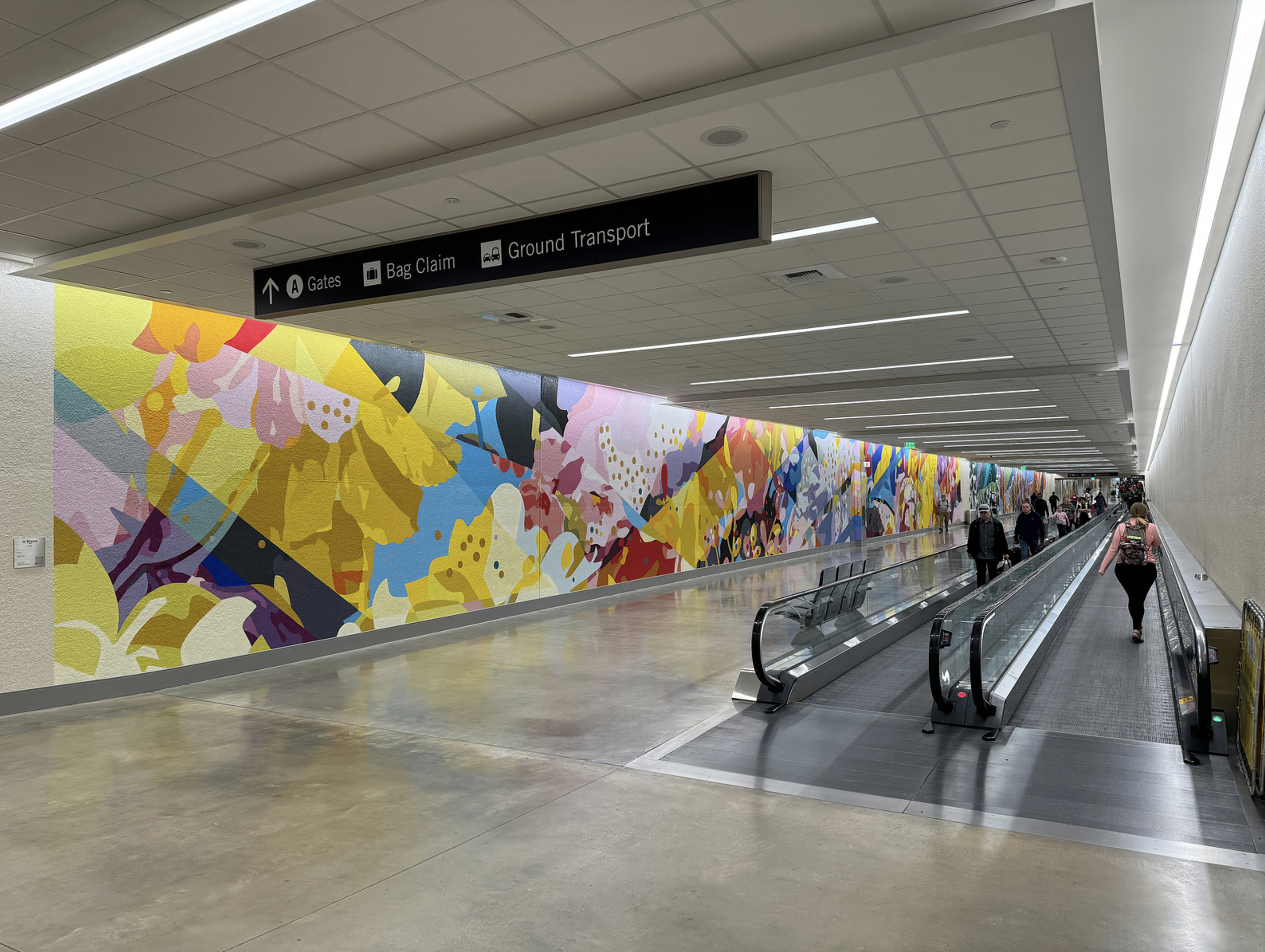 Work by the artist DAAS, Airport corridor with a colorful abstract mural on the left wall and a moving walkway on the right, with travelers walking along it. Overhead sign points to gates, bag claim, and ground transport.