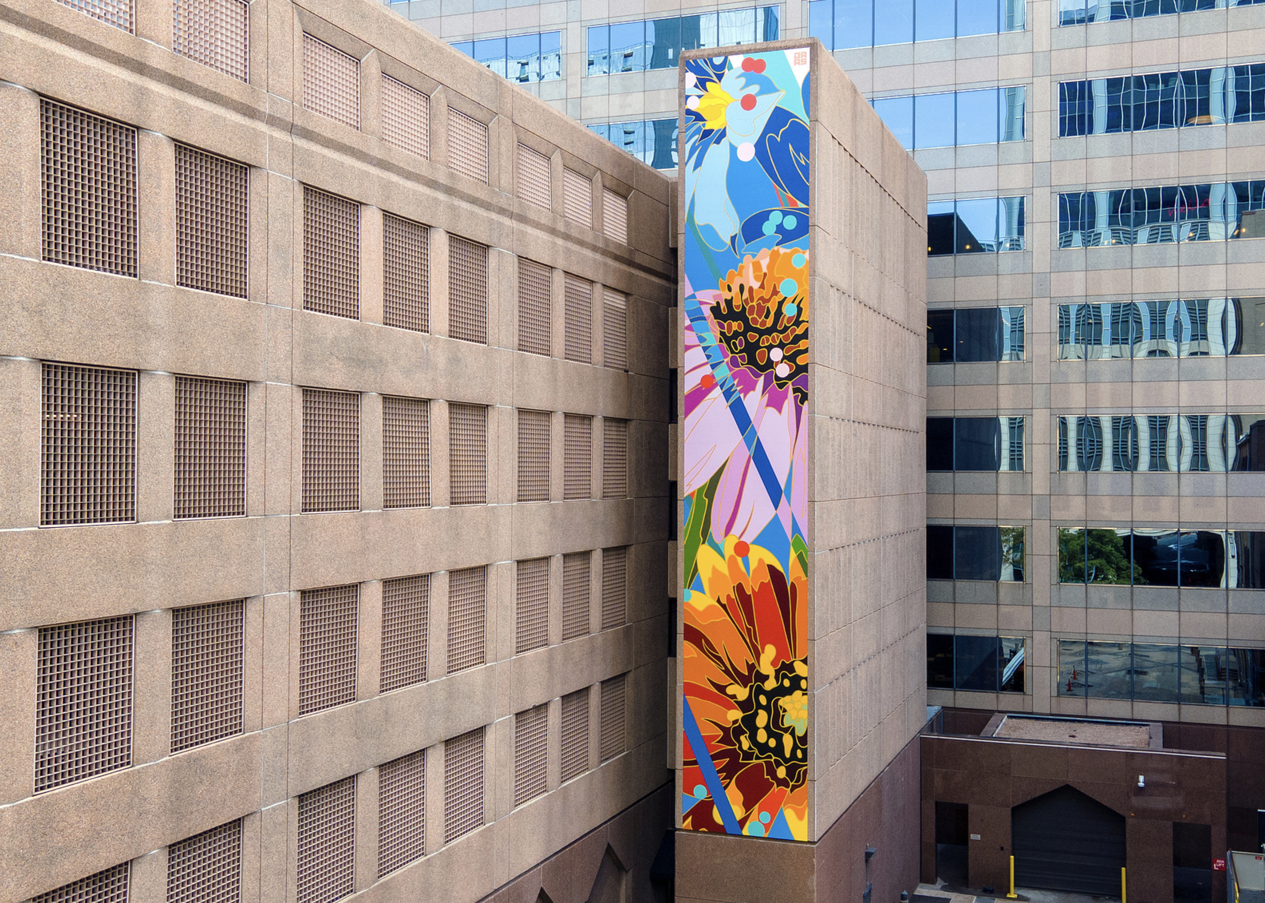 Work by the artist DAAS, A tall, colorful mural on the side of a building, featuring abstract floral and natural designs with bold colors, located between two beige and glass office buildings.