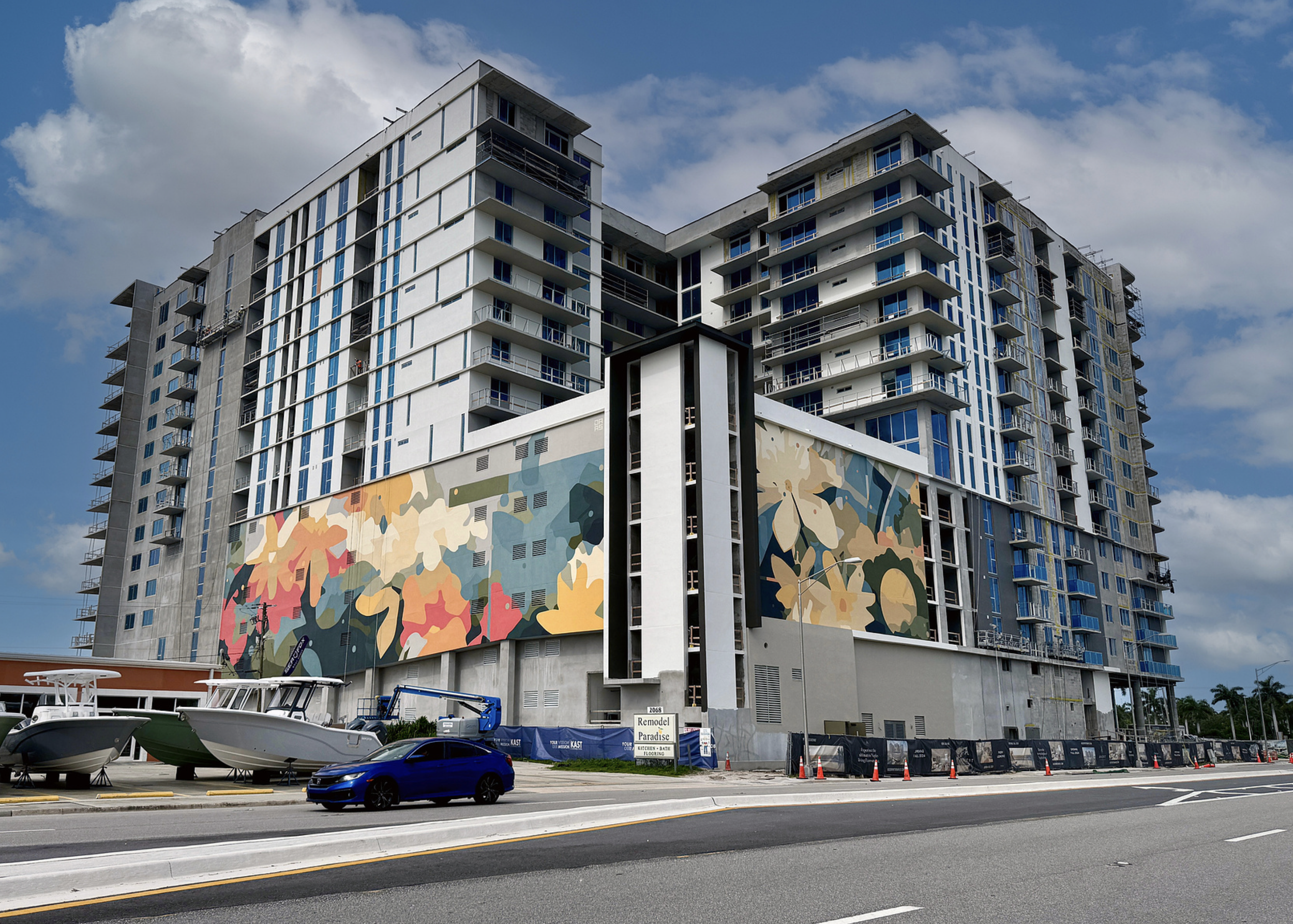 Work by the artist DAAS, Under construction multi-story residential building with colorful mural on the lower level, parked boats, and a blue car in the foreground under a partly cloudy sky.