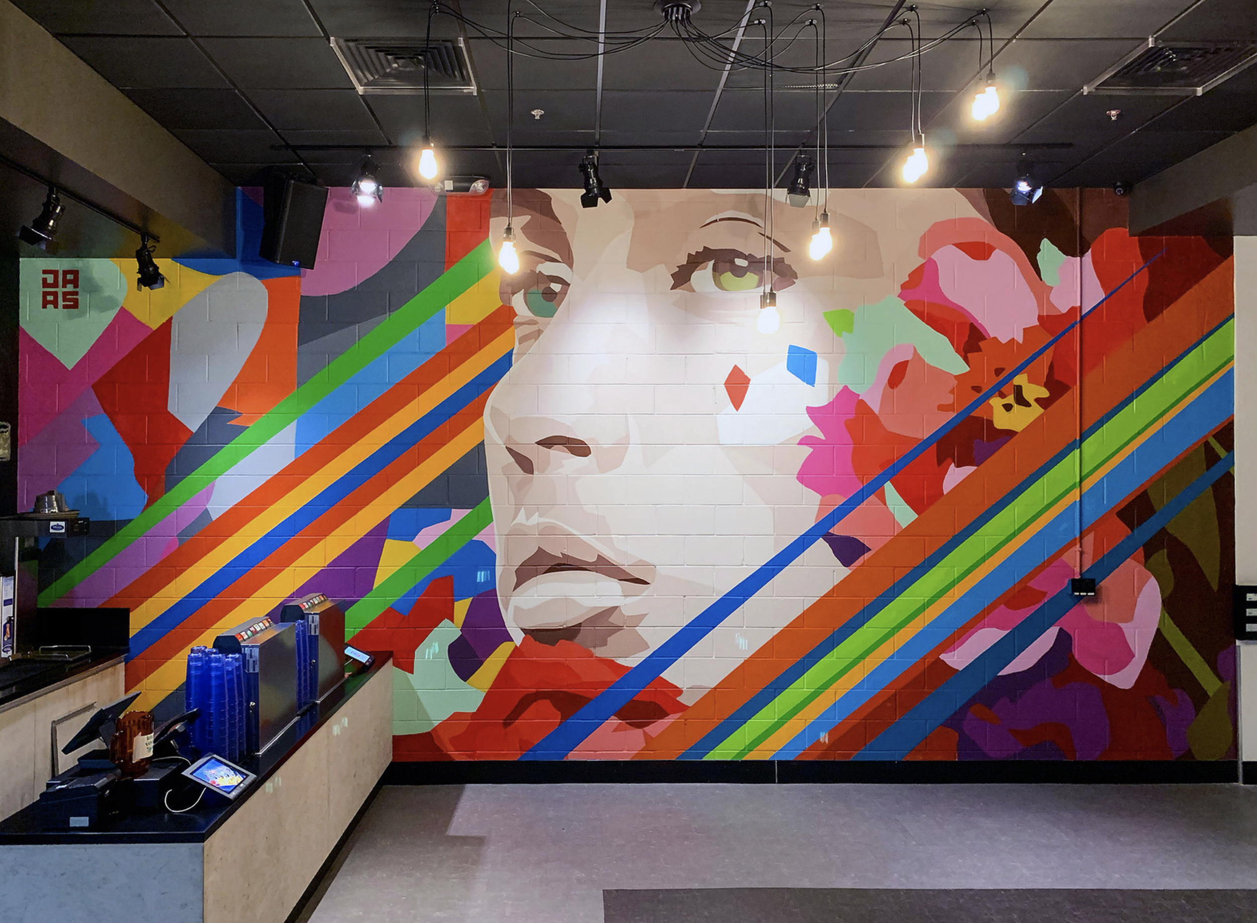 Work by the artist DAAS, Colorful mural of a woman's face with vibrant geometric patterns and rainbow stripes on a brick wall inside a modern cafe or lobby.