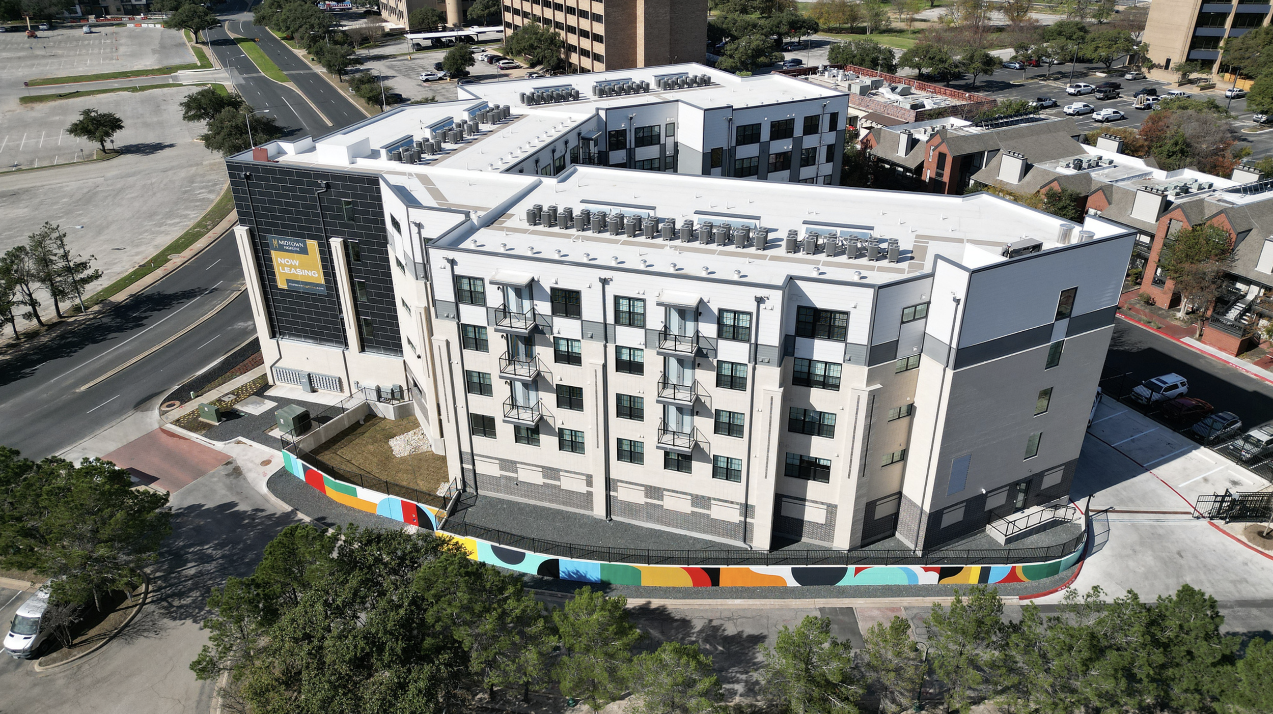 Work by the artist DAAS, A modern multi-story apartment building with a rooftop HVAC system, balconies, and a colorful abstract mural on the lower fencing, situated in an urban area with parking lots and other residential buildings.