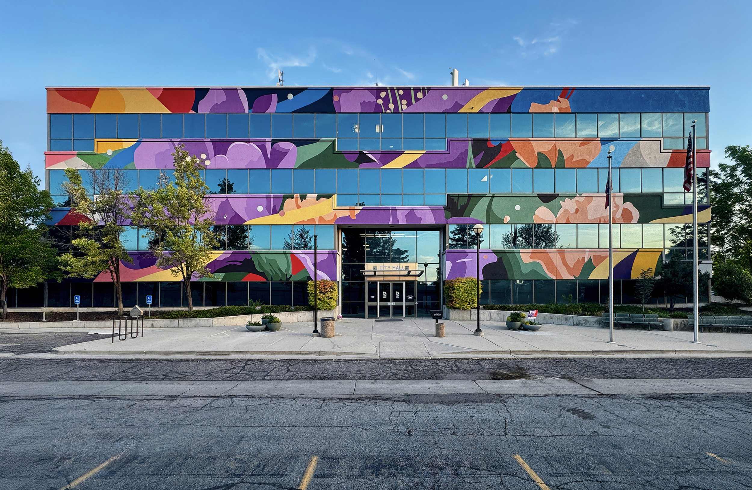 Work by the artist DAAS, Colorful mural on a glass building facade with trees and parking lot in front.