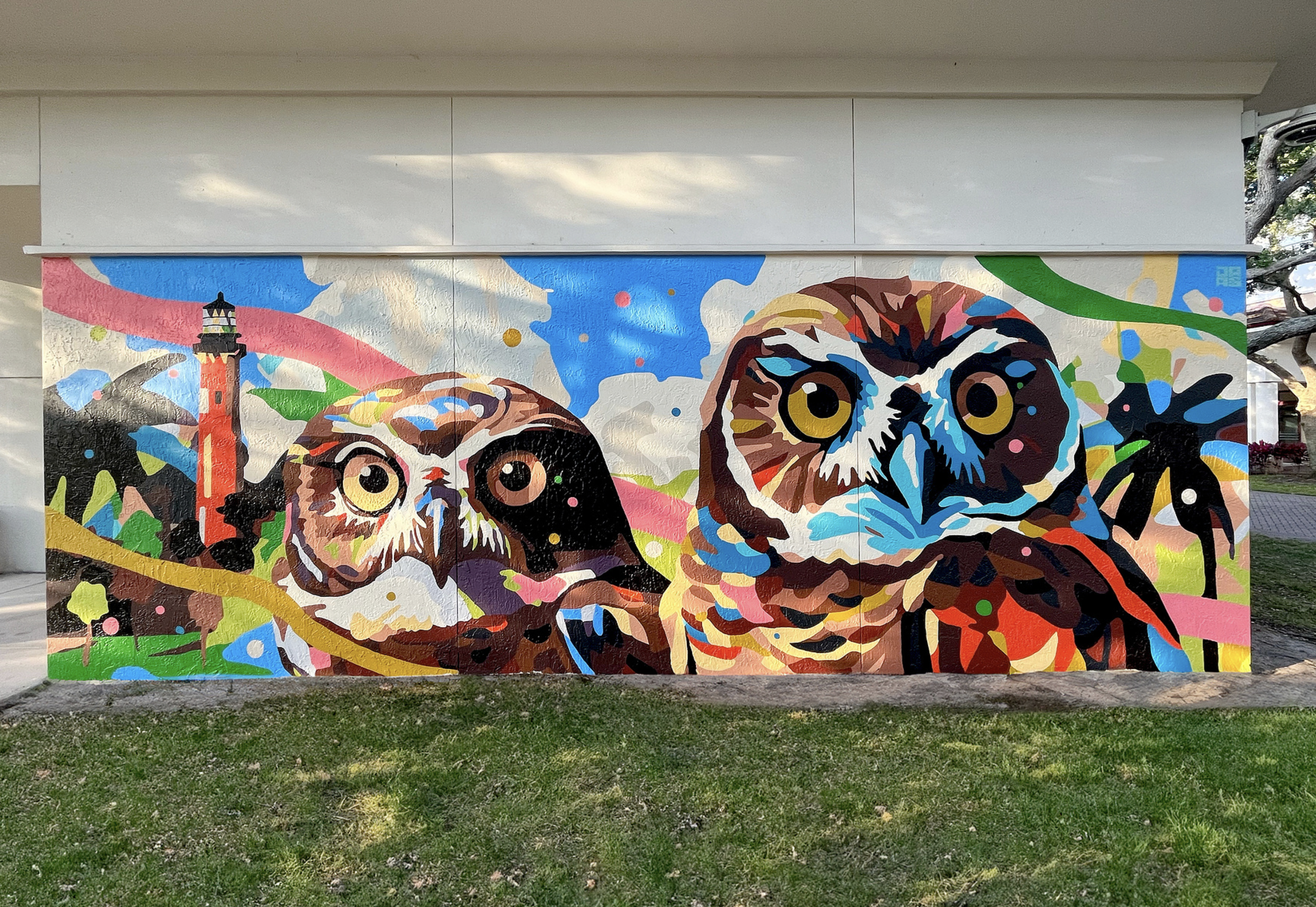Work by the artist DAAS, Colorful mural of two owls with a lighthouse, trees, and abstract shapes in the background.