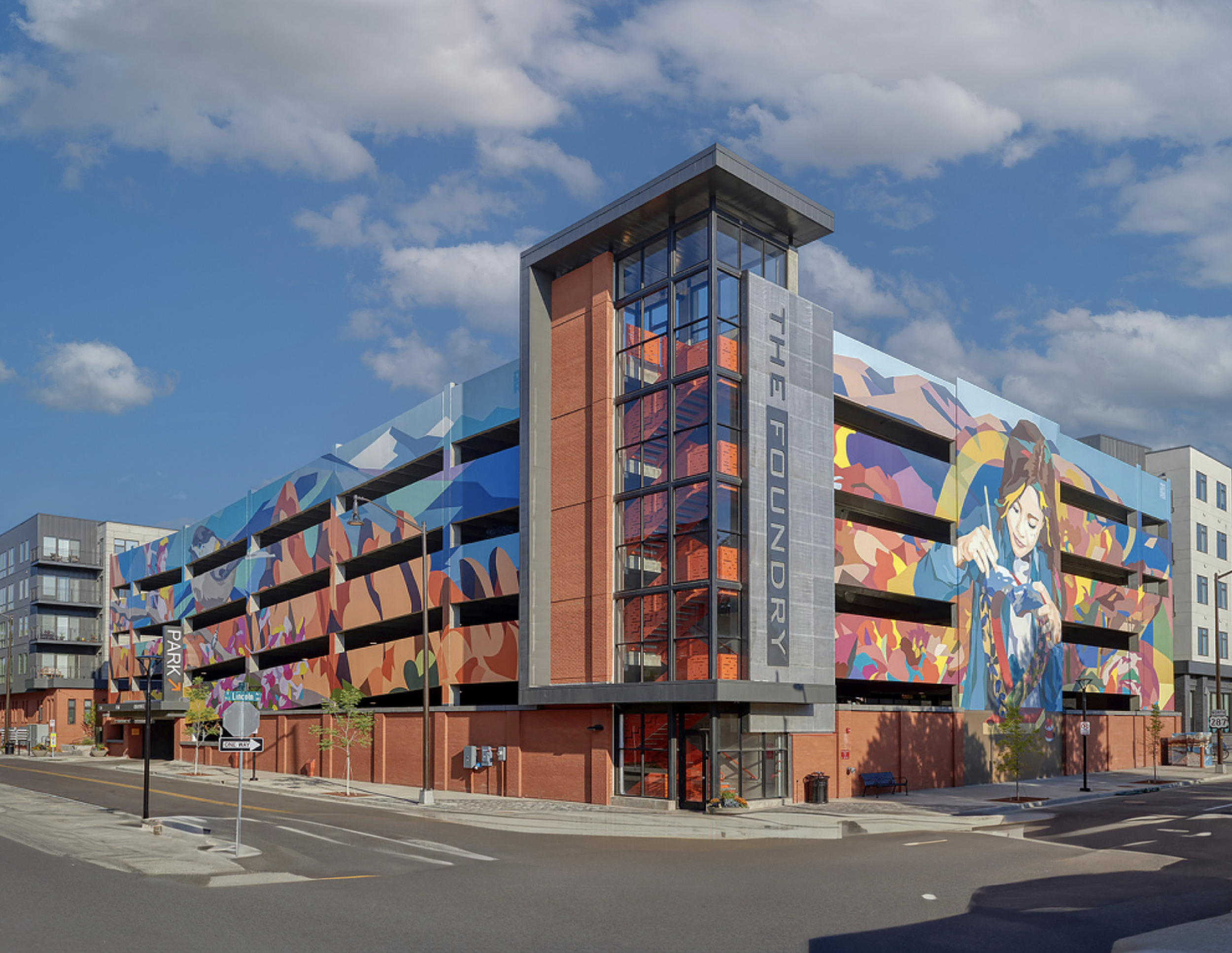 Work by the artist DAAS, A colorful, modern multi-story parking garage with a vibrant mural of a woman holding a camera on its side, located in an urban area under a blue sky with scattered clouds.