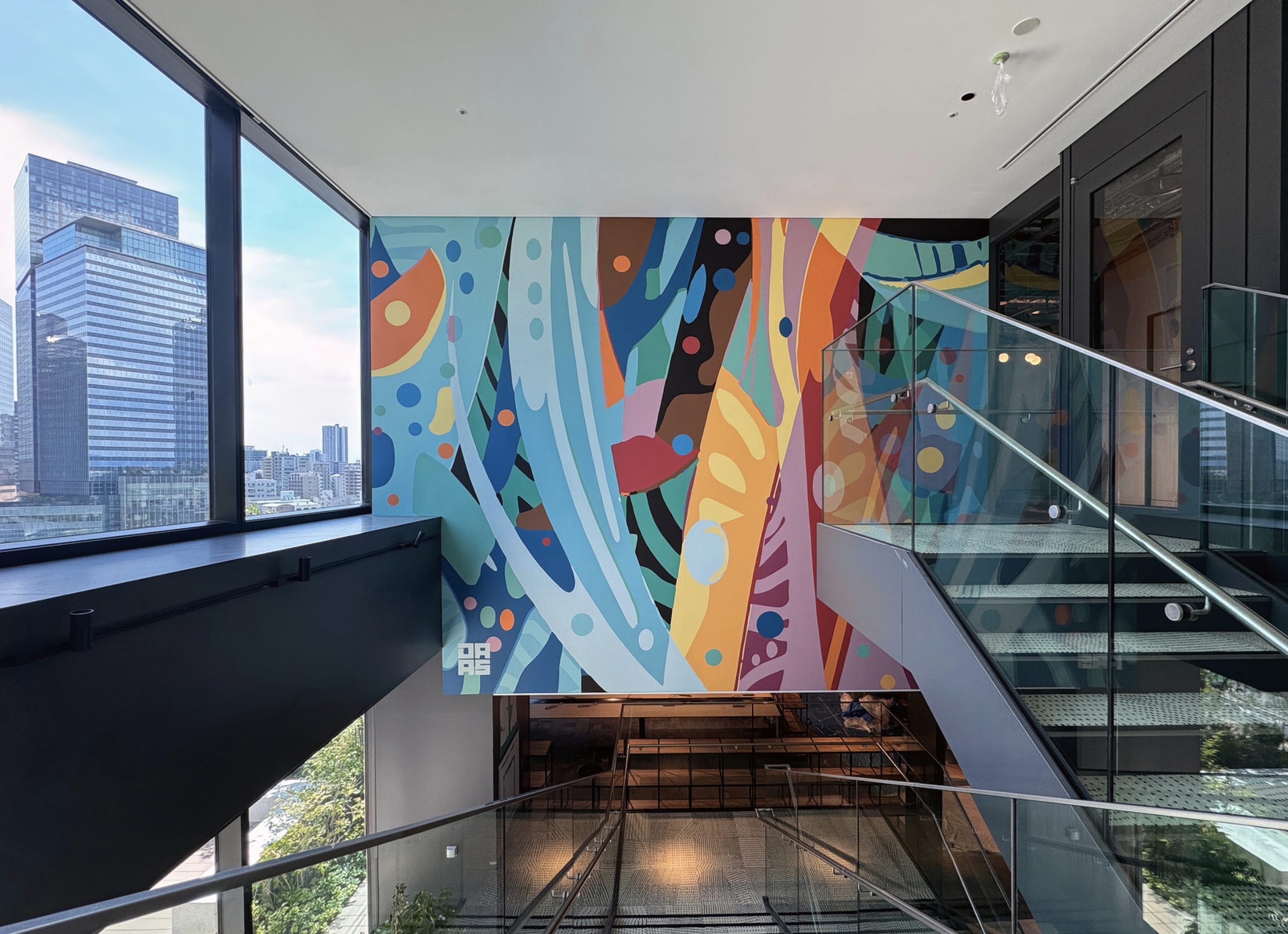 Work by the artist DAAS, Interior of a modern building with large windows, a colorful abstract mural on the wall, glass staircase, and city skyline visible outside.