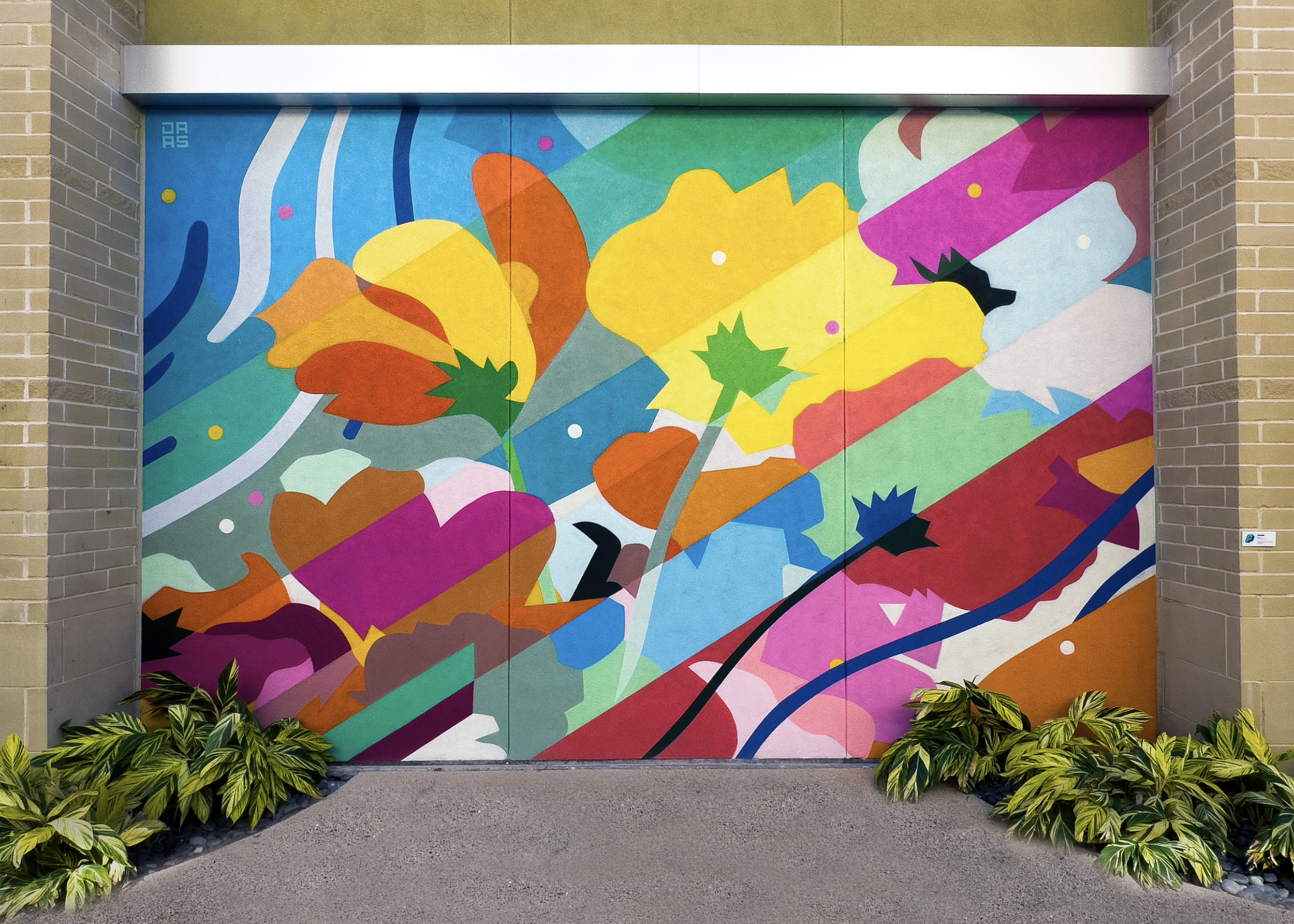 Work by the artist DAAS, Colorful abstract mural of flowers, leaves, and shapes painted on a garage door, with brick walls on either side and green plants at the bottom.