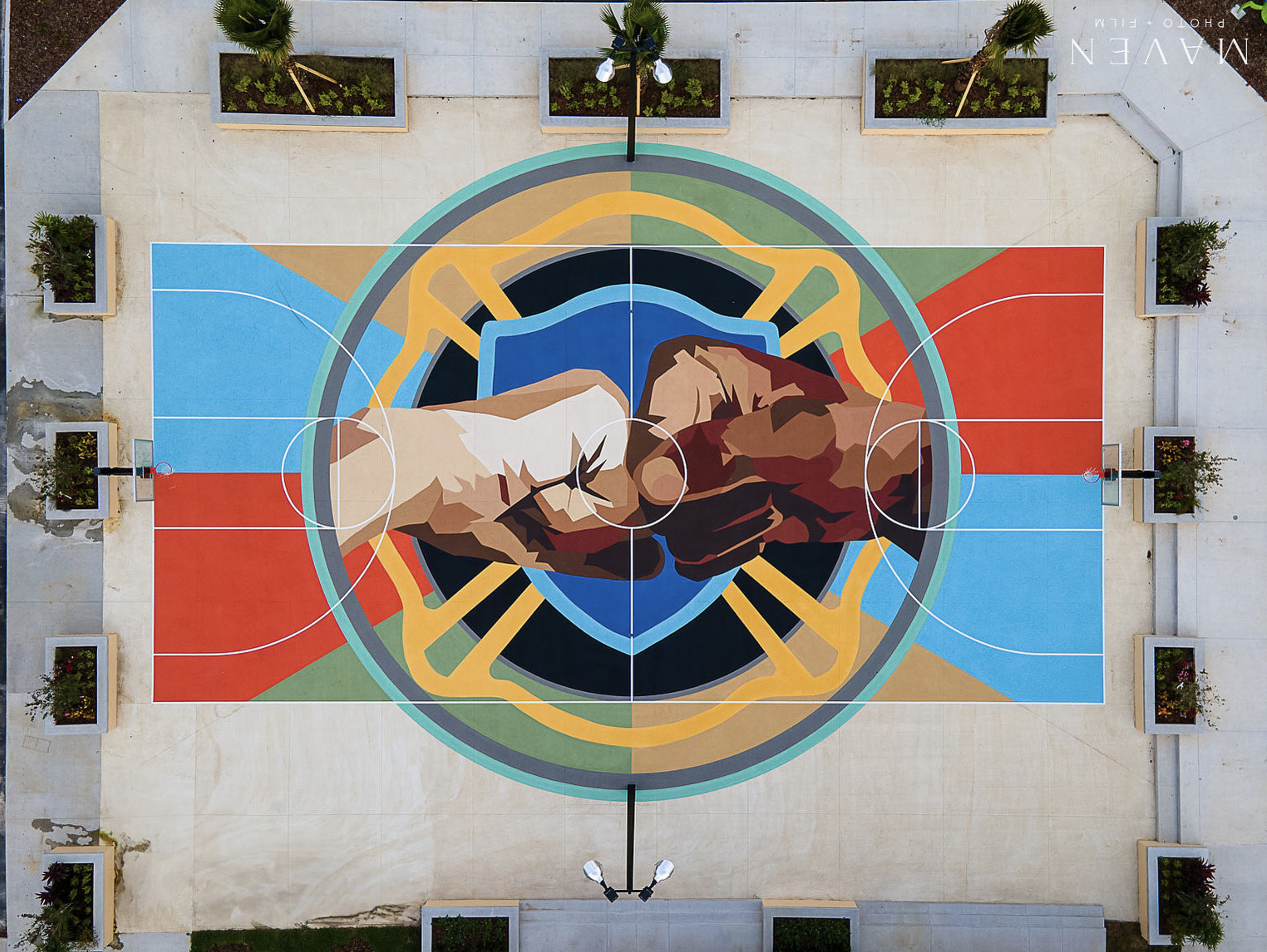 Work by the artist DAAS, A colorful mural on a basketball court features two hands engaging in a fist bump, set against a vibrant geometric background. The court is outdoors with potted plants surrounding it.