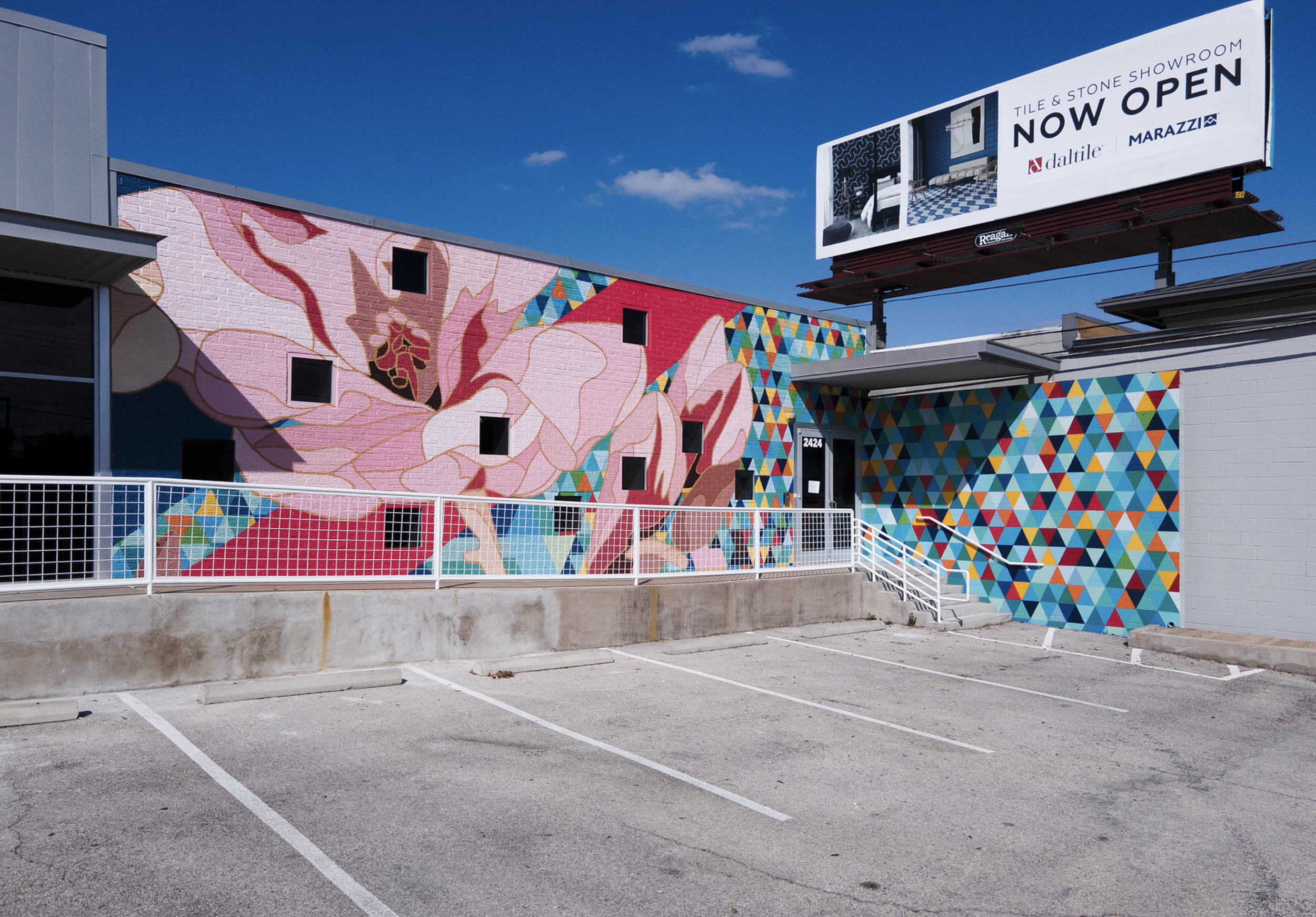 Work by the artist DAAS, Colorful mural of a large pink flower painted on the side of a building in an empty parking lot, with a billboard sign on top of the building advertising a tile and stone showroom that is now open.