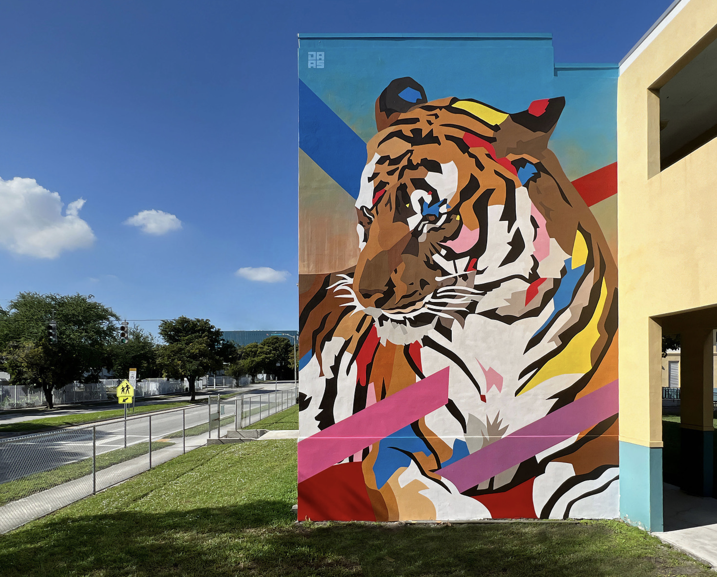 Work by the artist DAAS, Colorful mural of a tiger with abstract geometric shapes on a tall building exterior, in a grassy area beside a street.