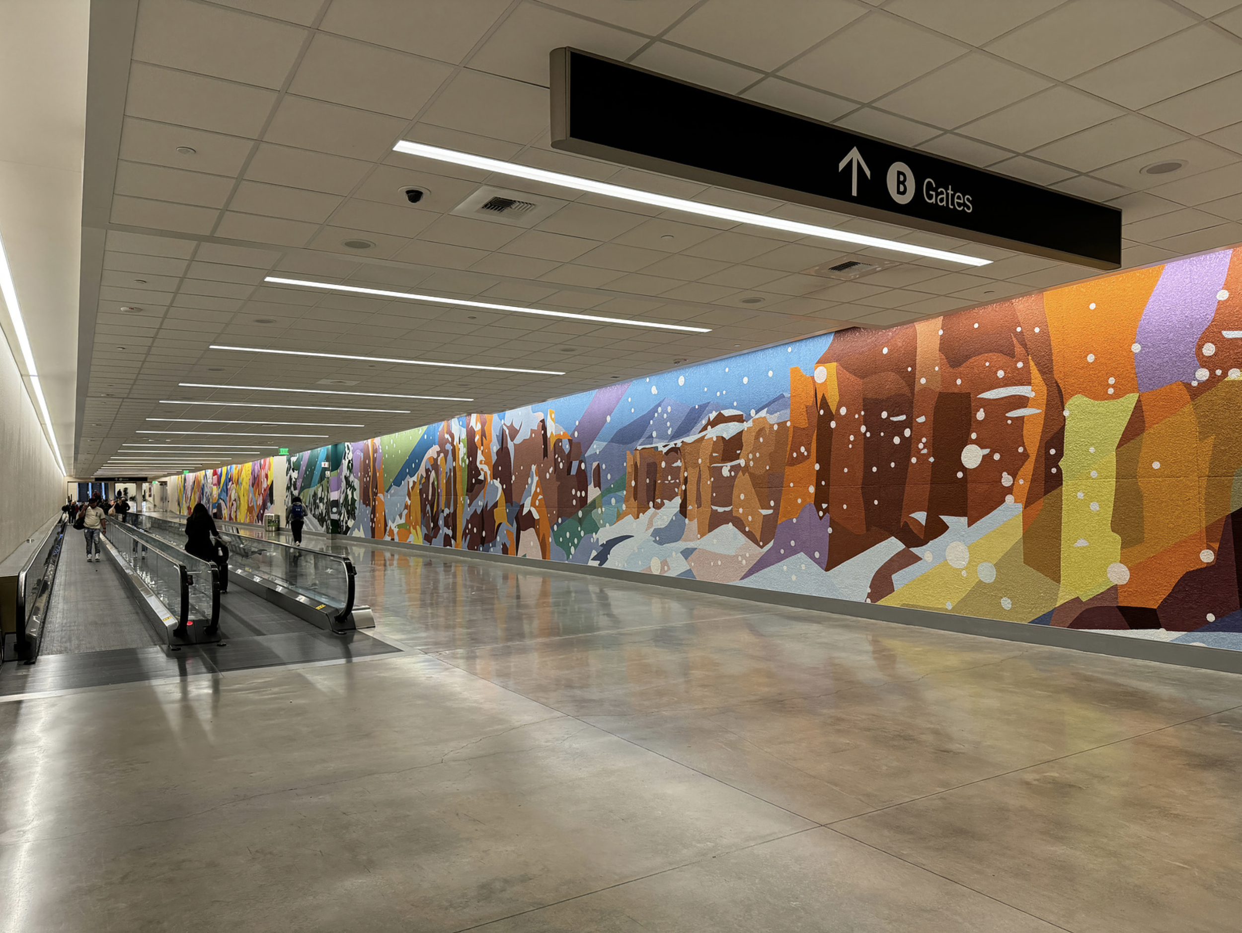 Work by the artist DAAS, An airport corridor with a colorful abstract mural on the right wall, moving walkways on the left, and a sign overhead indicating Gates B ahead.