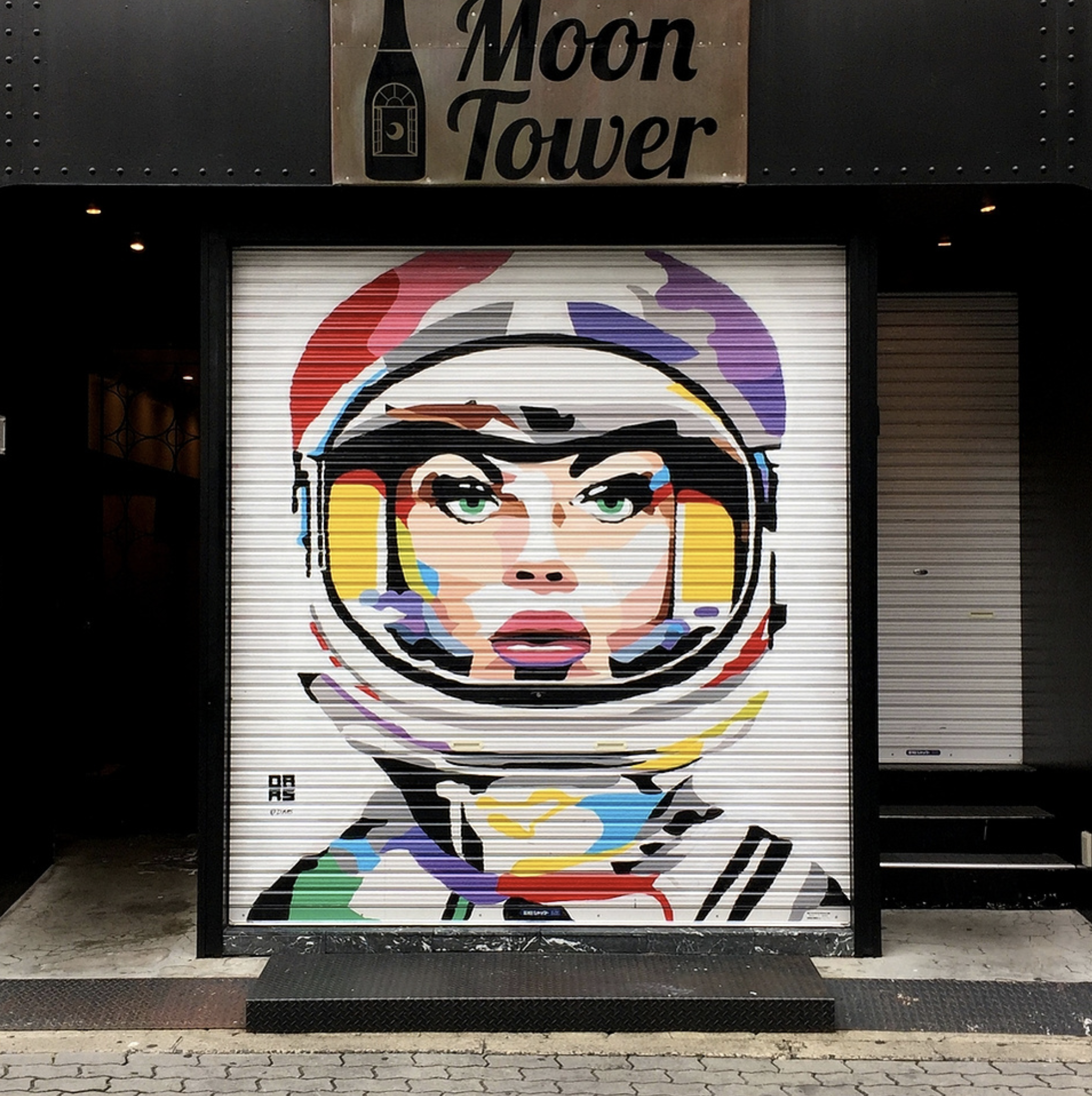 Work by the artist DAAS, Colorful mural of a female astronaut with striking green eyes, wearing a space helmet, painted on a metal rolling door in front of a business named 'Moon Tower'.