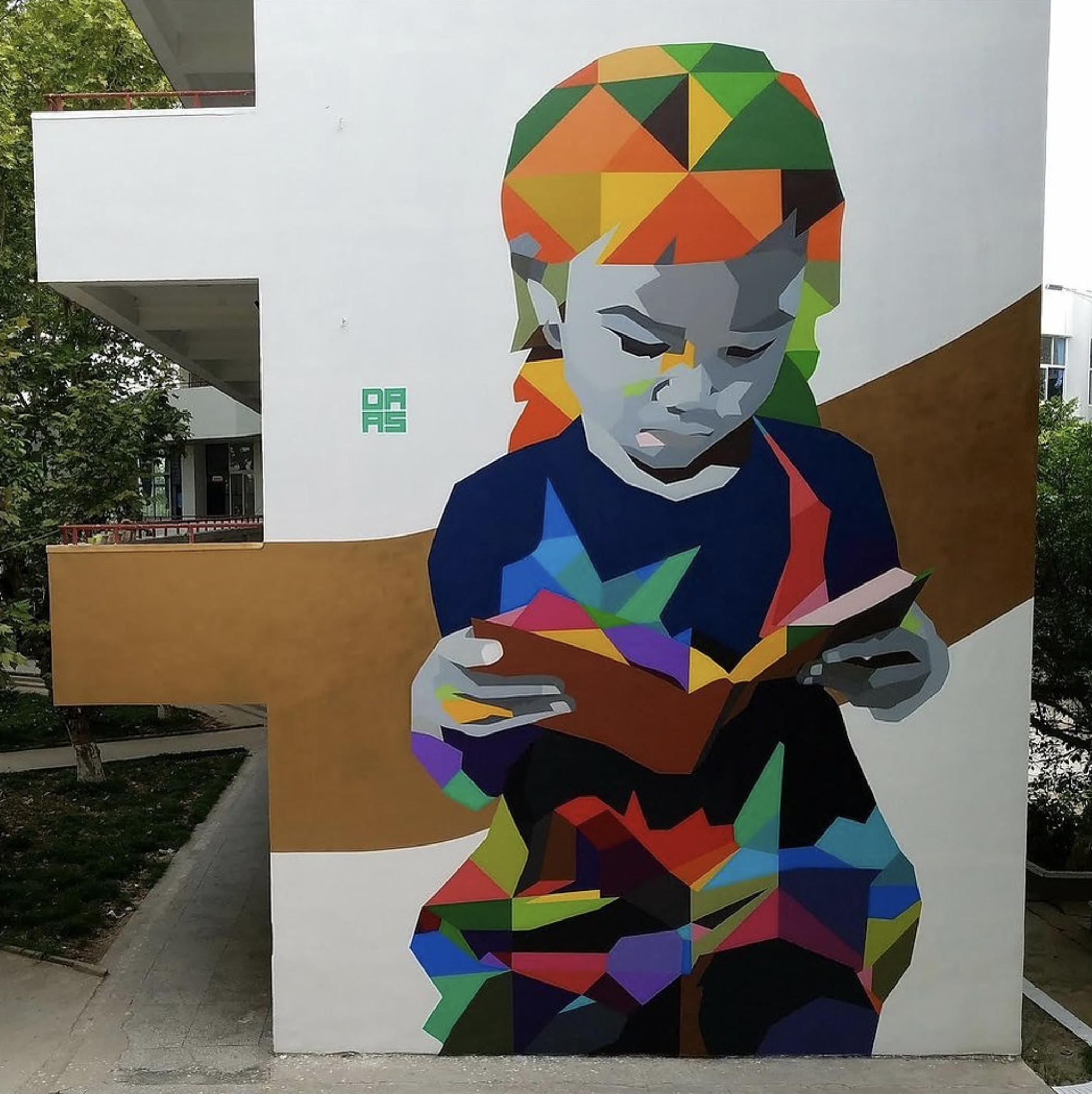 Work by the artist DAAS, Colorful geometric street art mural of a young child reading a book, on the side of a building with trees in the background.