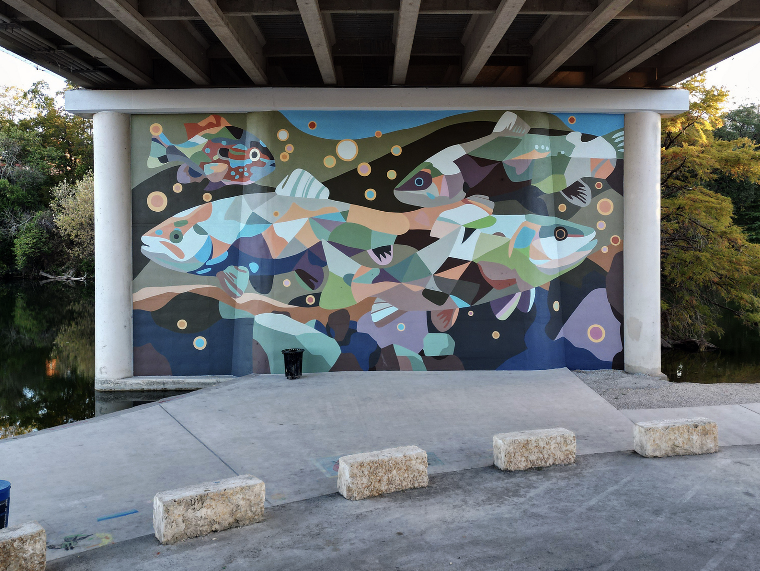 Work by the artist DAAS, A colorful mural of abstract fish and aquatic life painted under a bridge overlooking a body of water with trees in the background.