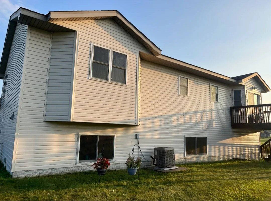 The side view of a two-story house with white vinyl siding, multiple windows, a small balcony, and a well-maintained grassy backyard. There are two potted plants and an air conditioning unit on the ground near the house.