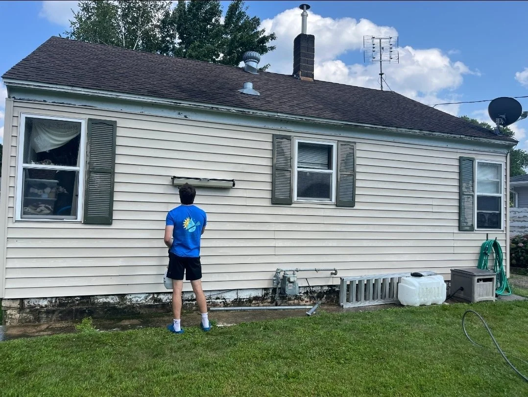 A person standing in front of the side of a beige house with three windows, green shutters, a brown roof, and various outdoor items on the ground, including a white water container, a ladder, and a green hose.