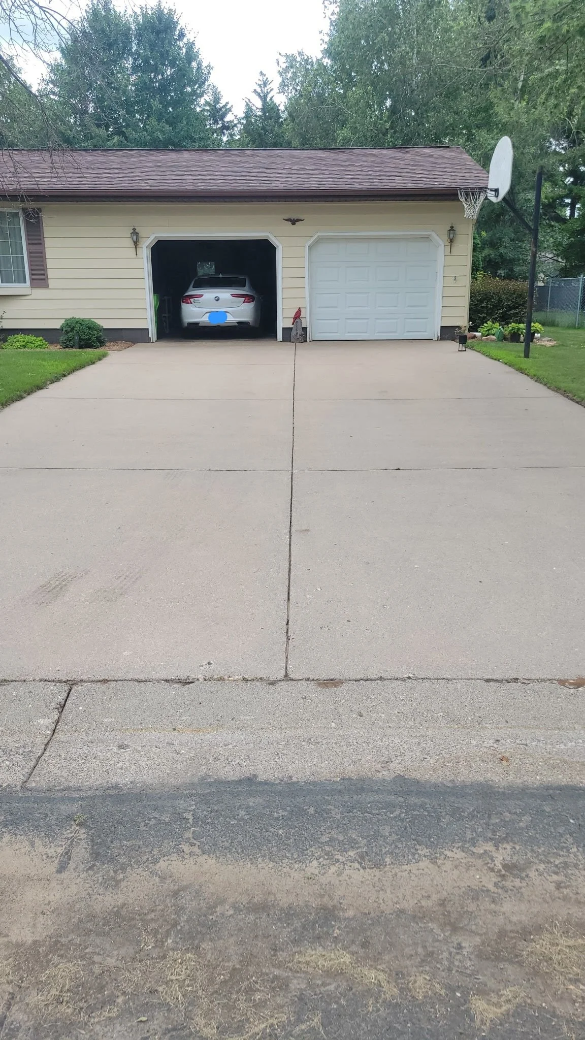 A house with a driveway, two garage doors, a parked car inside one garage, two outdoor lights on either side of the garage doors, a basketball hoop attached to the house, and a landscaped yard.