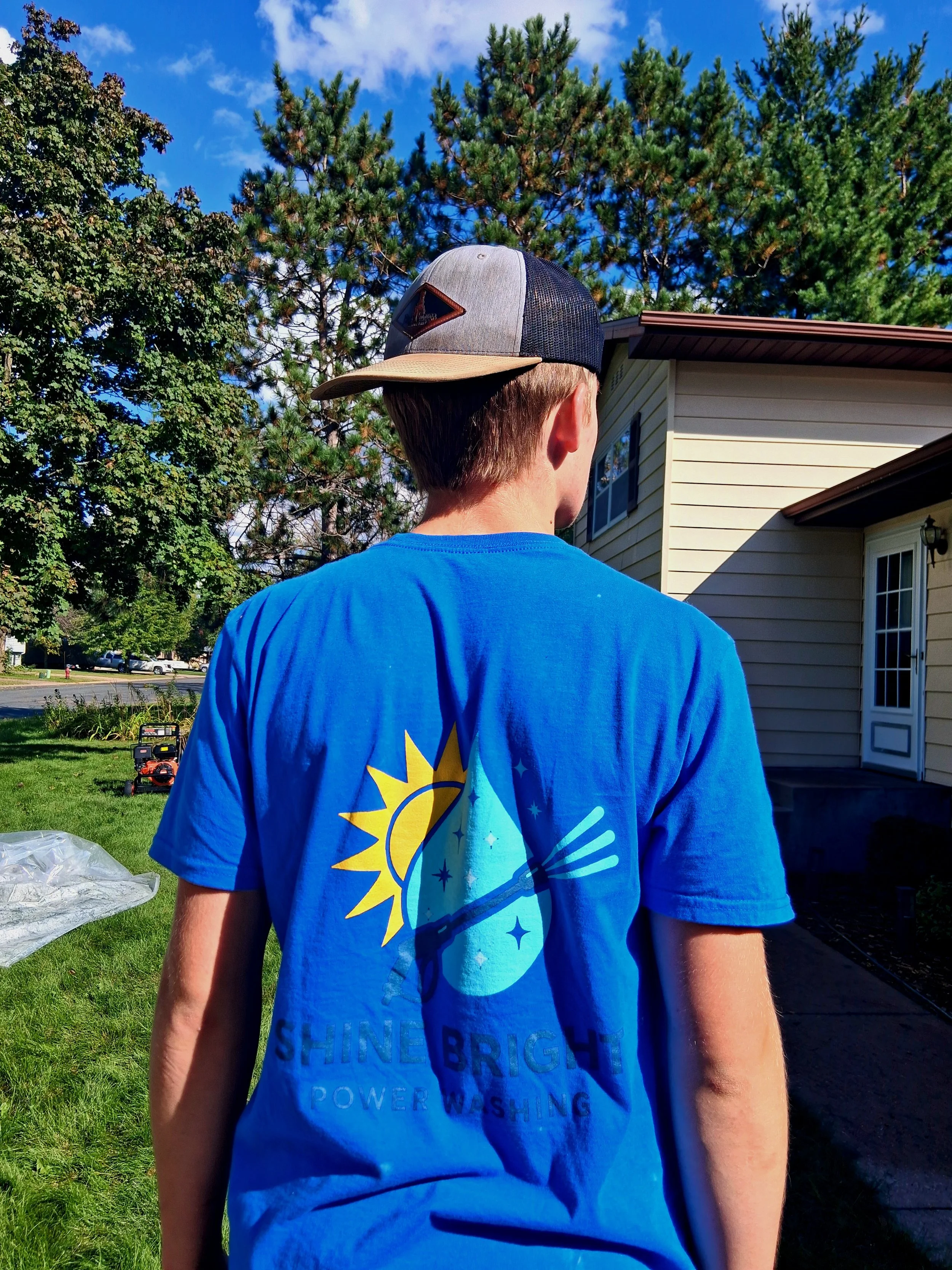 Back of a person wearing a blue t-shirt and a cap, standing outside near a house with trees and a blue sky in the background.