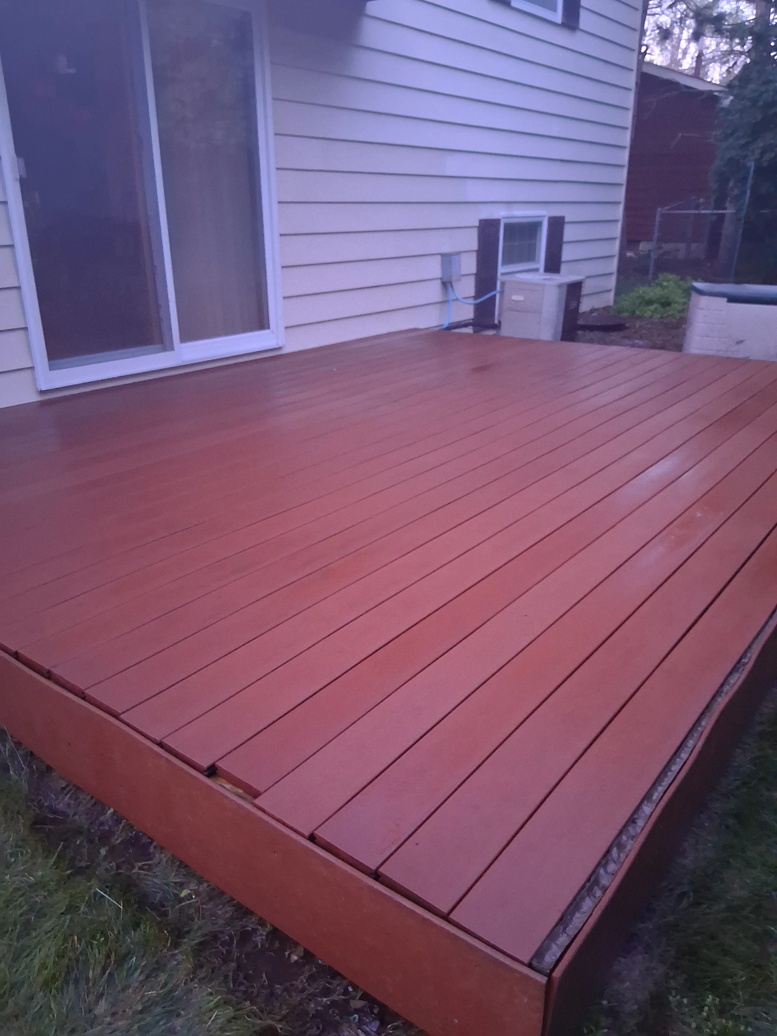 Newly built wooden deck outside a house, with beige siding, sliding glass door, and small windows.