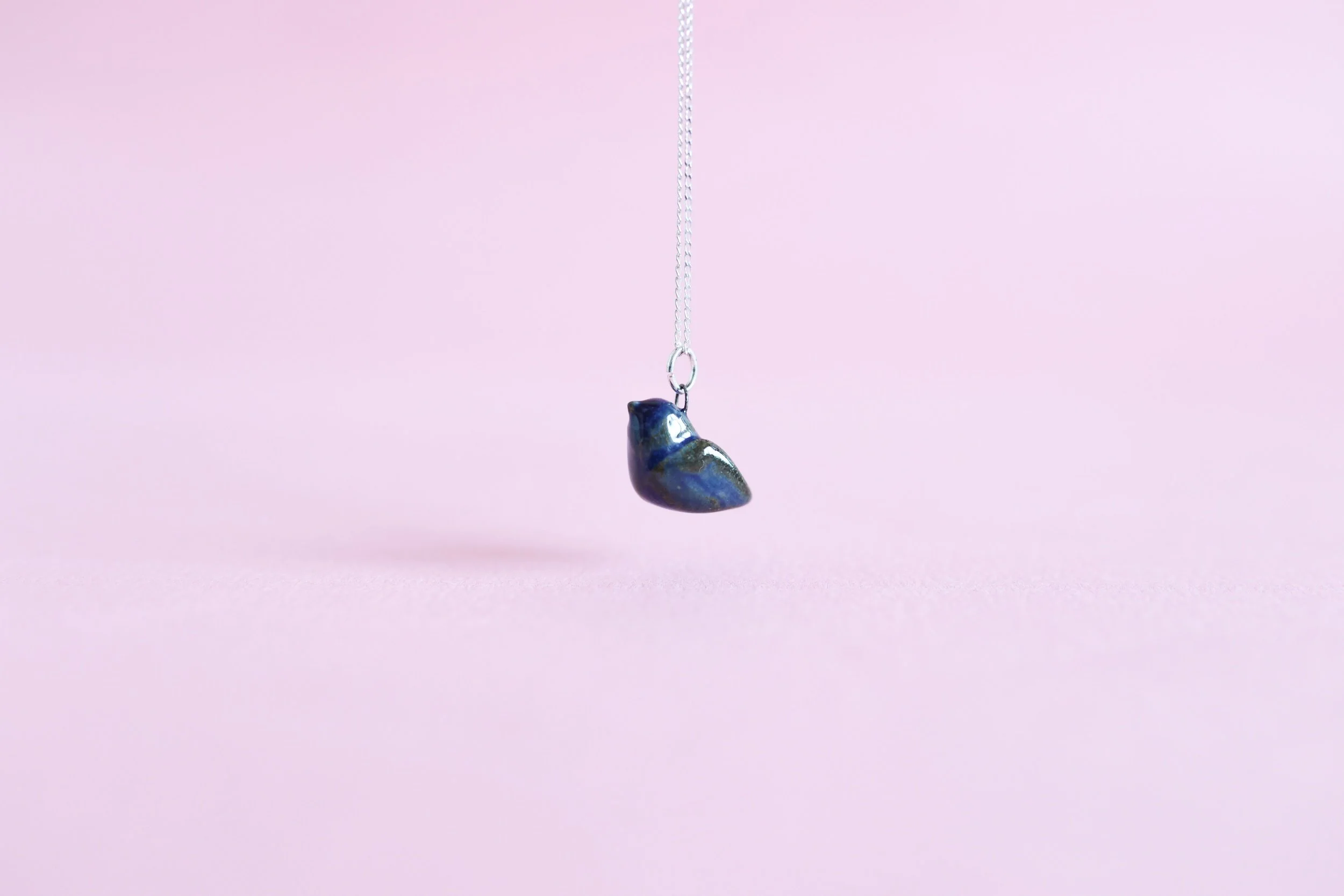 A navy blue ceramic bird is suspended in the air on a sterling silver chain in front of a pink background.