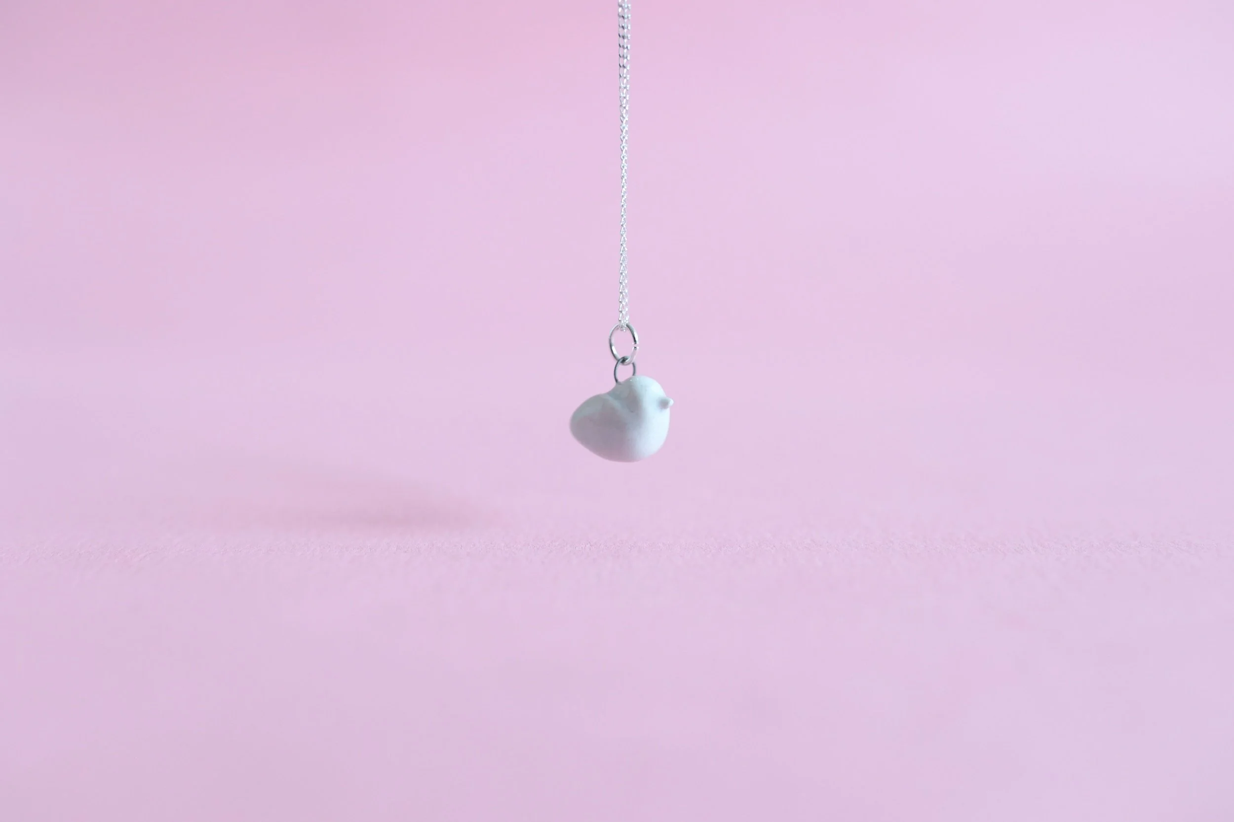 A turquoise ceramic bird is suspended in the air on a sterling silver chain in front of a pink background.