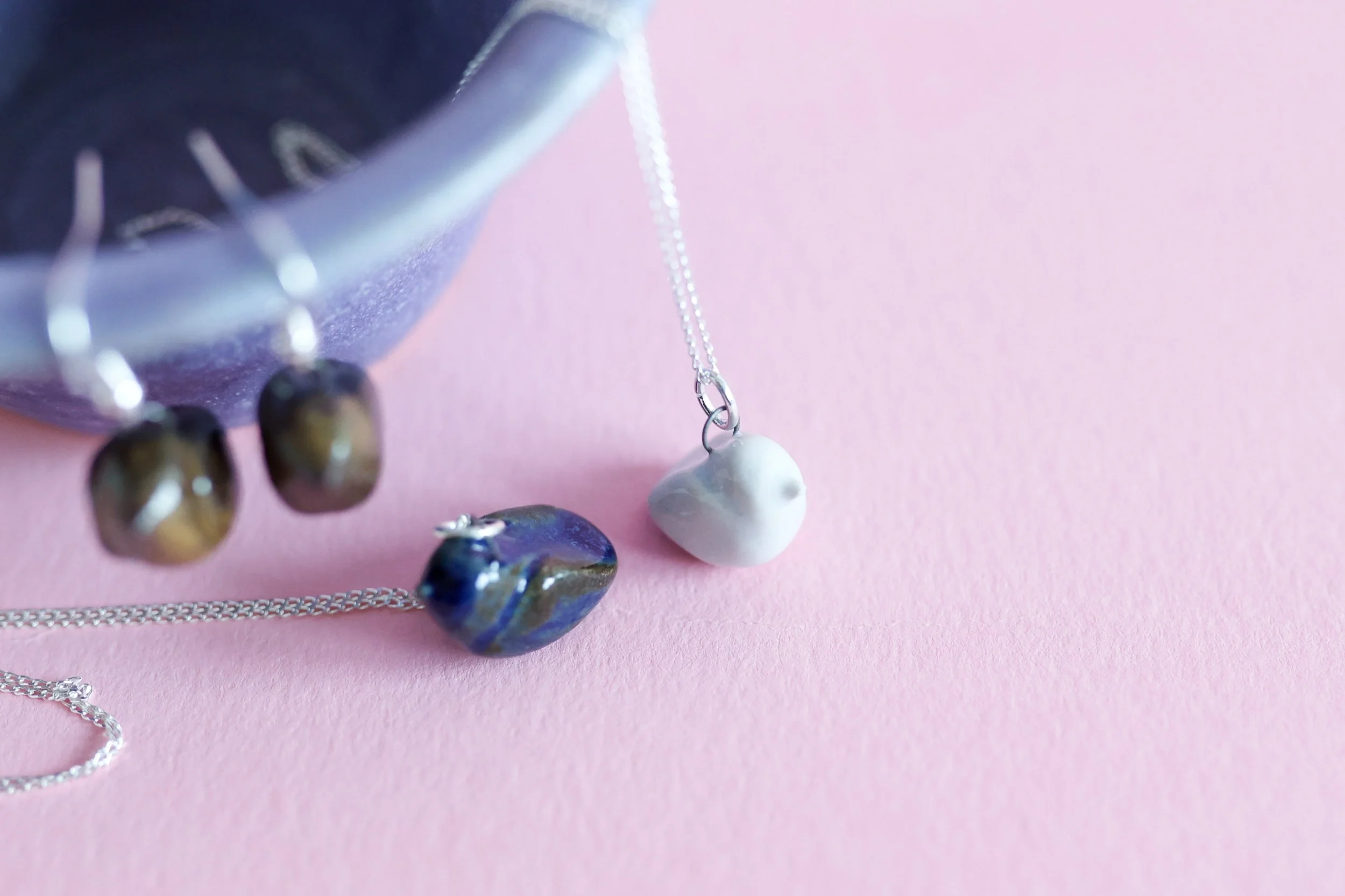 Small ceramic bird pendants, sitting on a pink background, there is also a purple dish in the image that has some brown bird earrings hanging from it.