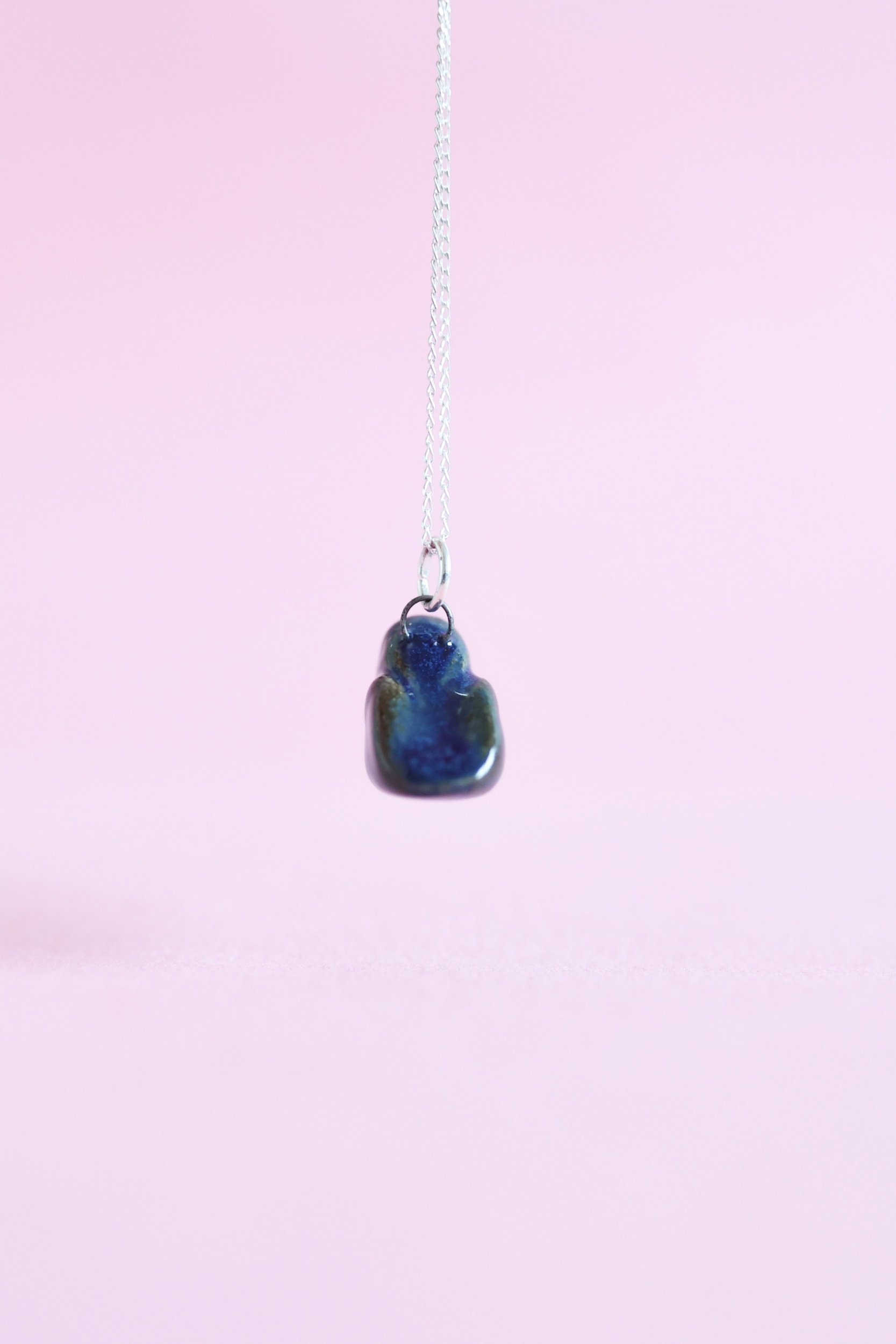A navy blue ceramic bird is suspended in the air on a sterling silver chain in front of a pink background.