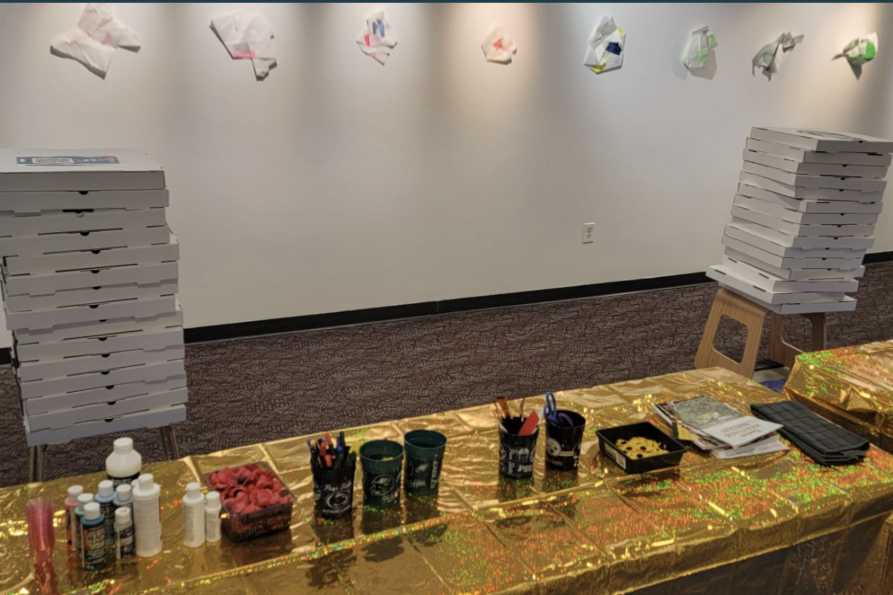 A table covered with a shiny gold tablecloth holds cups, markers, a container of pink foam, and papers. In the background, there are many stacked pizza boxes on two stools. The wall behind is decorated with paper butterflies.