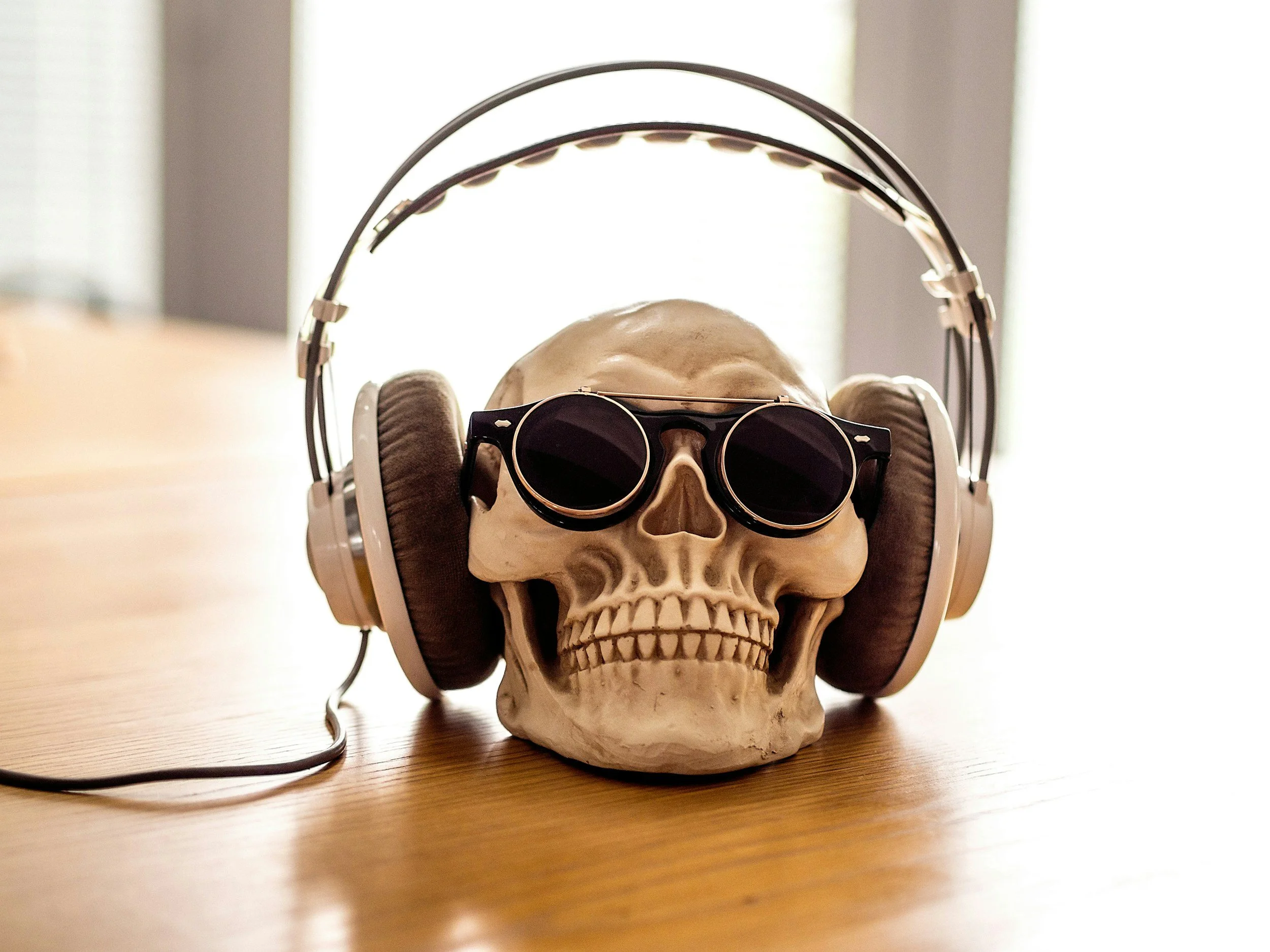 A skull wearing black sunglasses, over-ear headphones, and a headset microphone, placed on a wooden surface with a bright, blurred background.