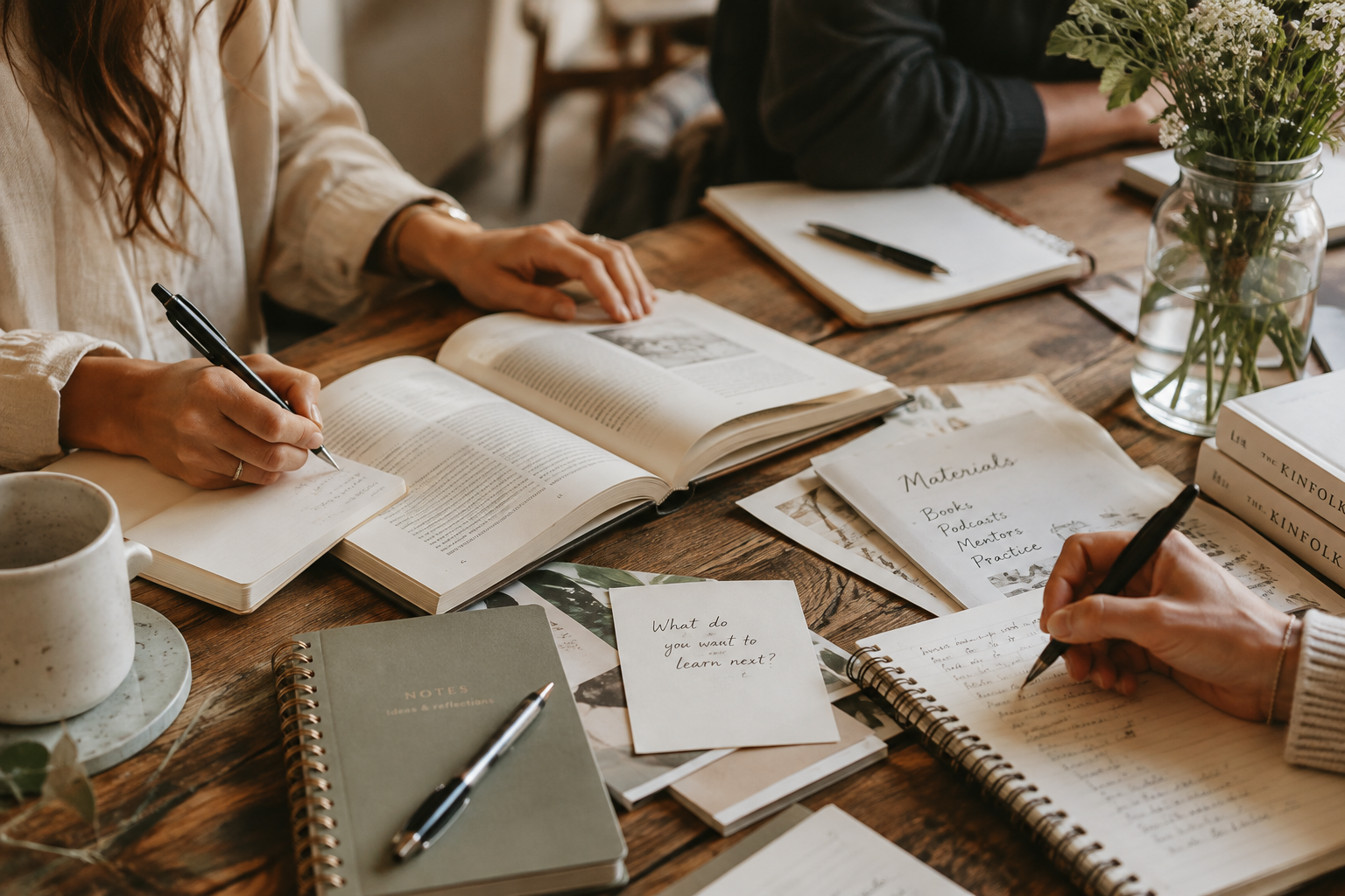 People working at a wooden table with open books, notebooks, notebooks, papers, pens, a mug, and a vase of flowers.