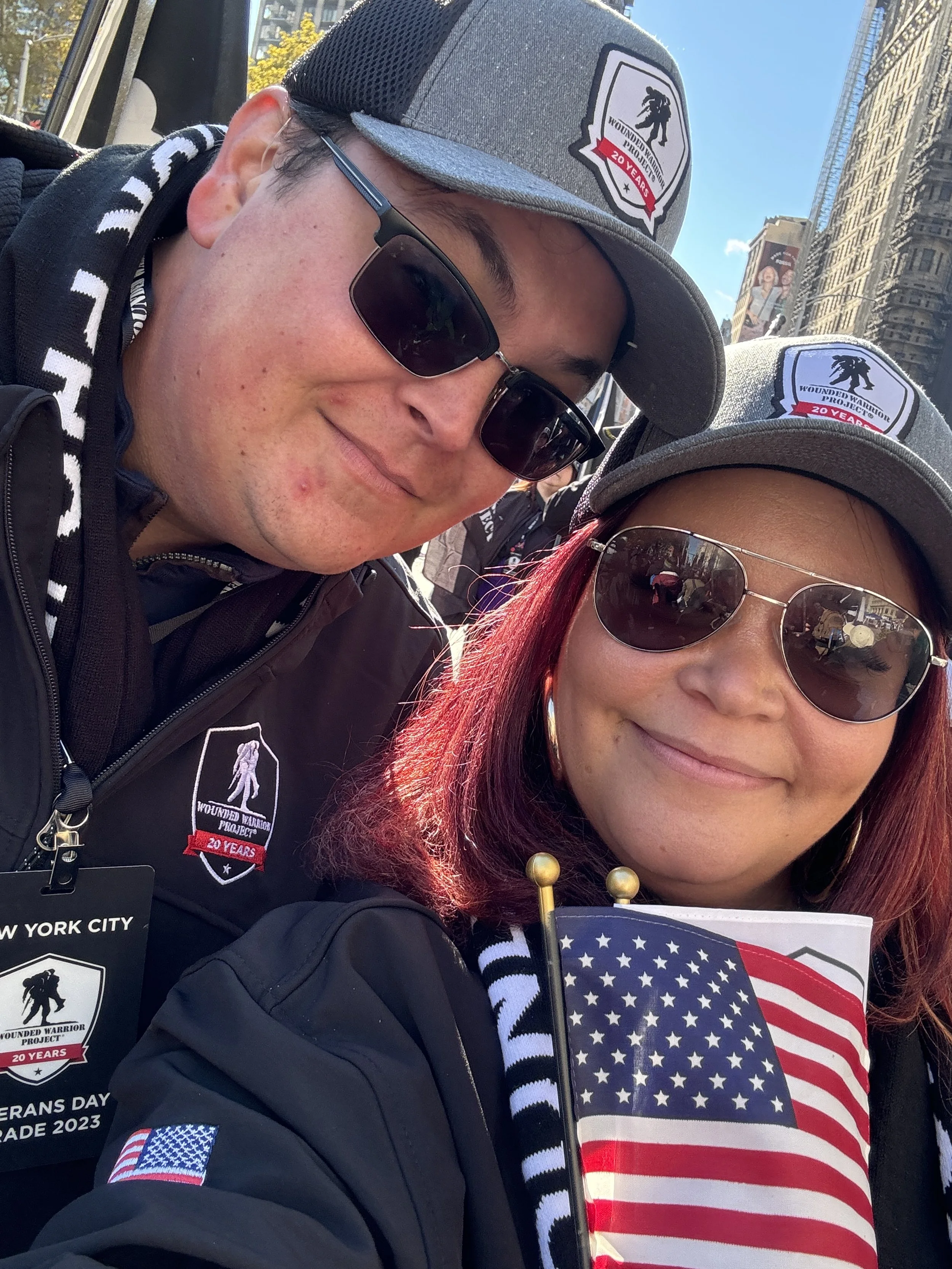 Nourich Founders,  Briana McCrae Carr & Frank Carr wearing WWP gray hats, sunglasses, and black jackets, smiling for a selfie in NYC for the Veterans Day Parade with buildings in the background. One of them is holding a small American flag.